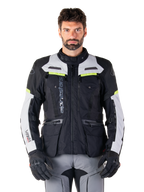 Man draagt een Alpinestars Bogotá Pro Drystar® Jas, Black Ice Gray en Yellow Fluo, all-season adventure touring motorjas, waterdicht ontwerp met meerdere zakken en verticale branding, gecombineerd met zwarte handschoenen en een grijze motorbroek