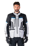 Alpinestars Bogota Pro Drystar jas, adventure touring motorjas, Ice Gray met zwarte en cyaanblauwe accenten, vooraanzicht met ventilatiepanelen en cargozakken, gedragen door een model met zwarte motorbroek en handschoenen