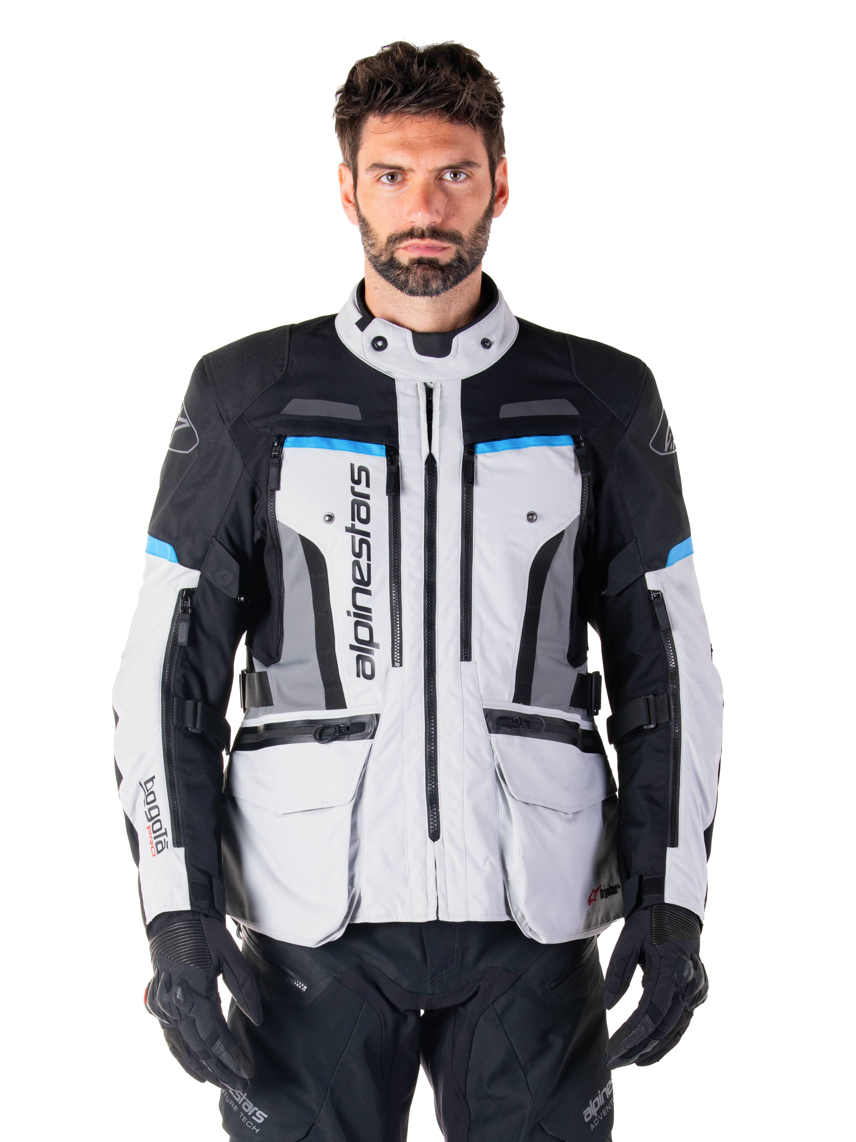 Alpinestars Bogota Pro Drystar jas, adventure touring motorjas, Ice Gray met zwarte en cyaanblauwe accenten, vooraanzicht met ventilatiepanelen en cargozakken, gedragen door een model met zwarte motorbroek en handschoenen
