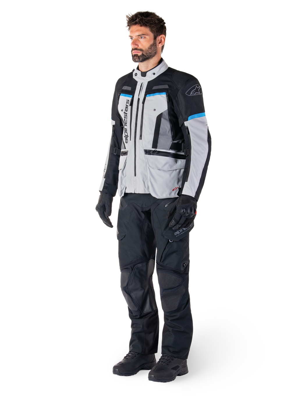 Mannelijke motorrijder die de Alpinestars Bogotá Pro Drystar® Jas draagt in Ice Gray, zwart en cyaan, gecombineerd met een zwarte technische motorbroek en handschoenen, adventure touring-uitrusting voor prestaties onder alle weersomstandigheden.