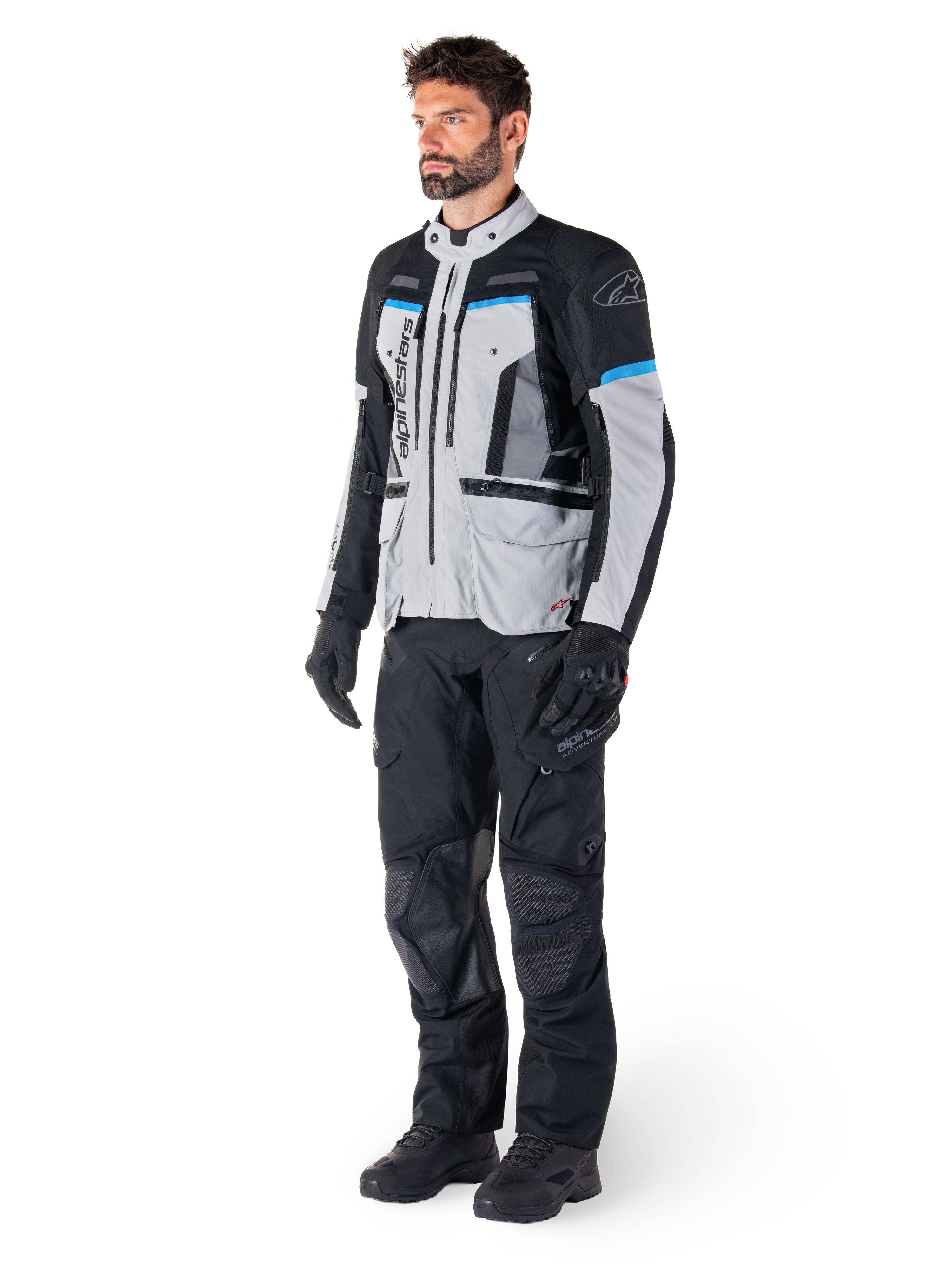 Mannelijke motorrijder die de Alpinestars Bogotá Pro Drystar® Jas draagt in Ice Gray, zwart en cyaan, gecombineerd met een zwarte technische motorbroek en handschoenen, adventure touring-uitrusting voor prestaties onder alle weersomstandigheden.