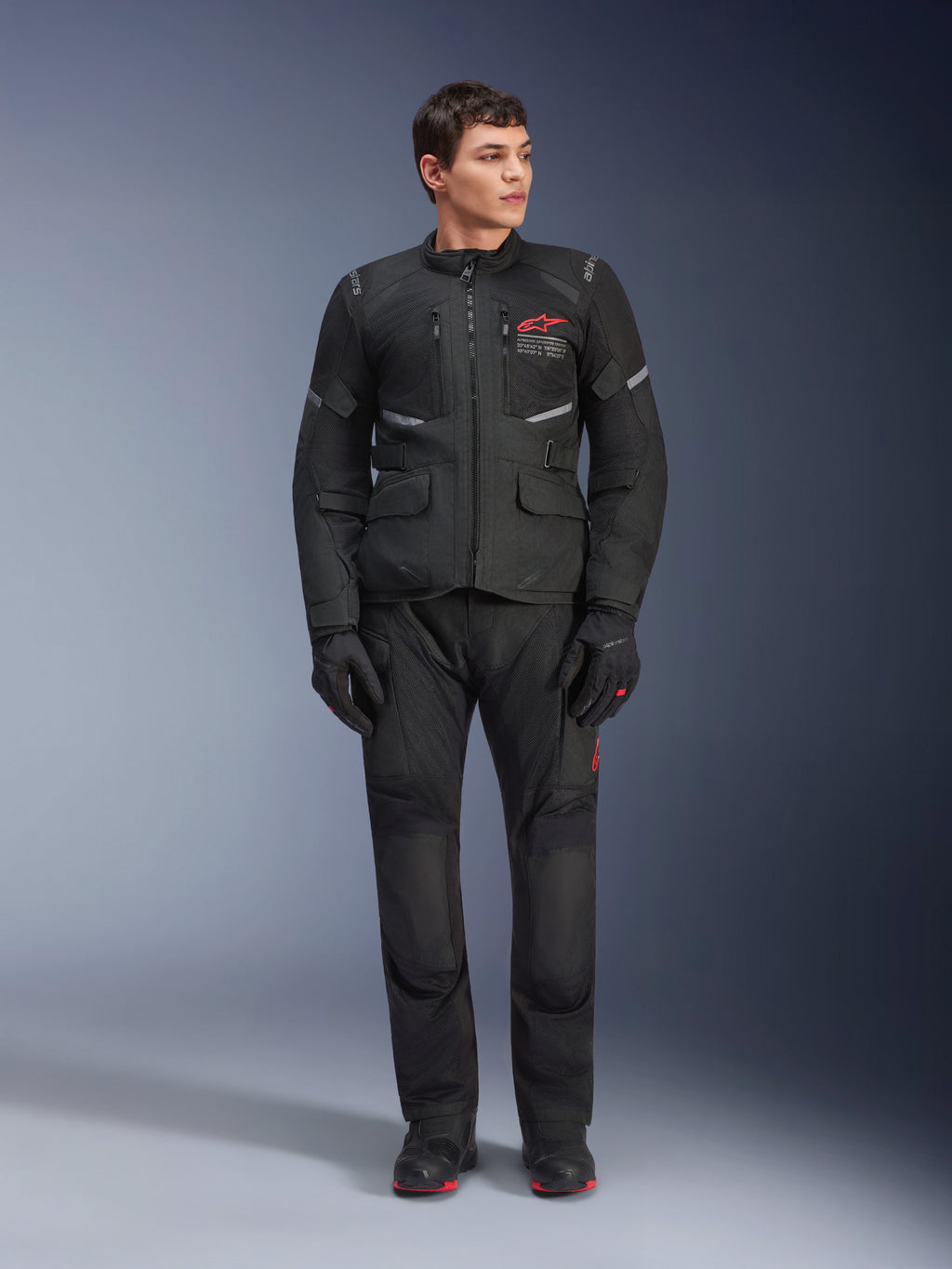 Mannelijk model in de Alpinestars Andes Air Drystar® Jas in het zwart, adventure touring motoruitrusting met bijpassende broek en handschoenen, voorzien van een rood logo op de borst, mesh-ventilatie en functionele zakken.