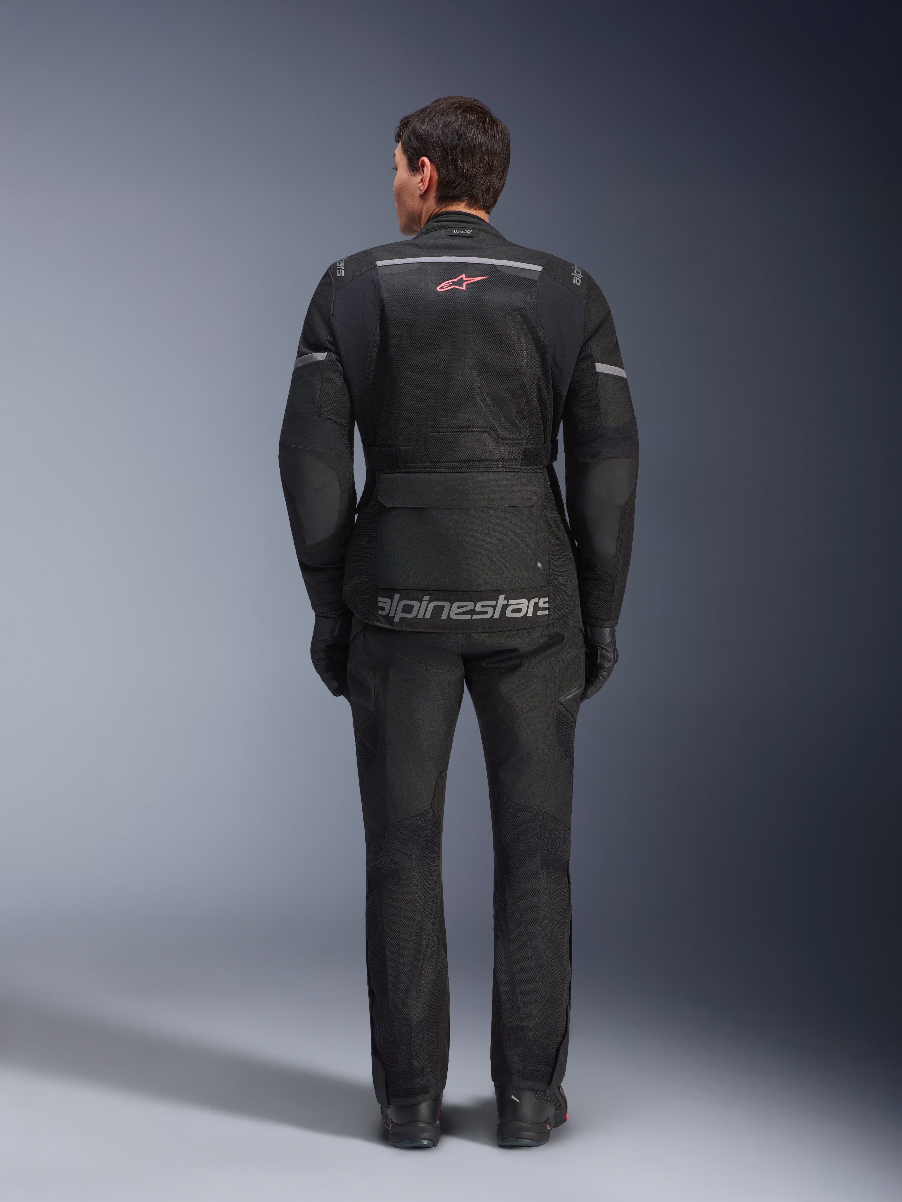 Achteraanzicht van een rijder die de Alpinestars Andes Air Drystar® Jas in het zwart draagt, voorzien van uitgebreide mesh ventilatiepanelen op de rug, een rood Alpinestars logo en reflecterende accenten, gecombineerd met een bijpassende motorbroek en handschoenen voor adventure touring.