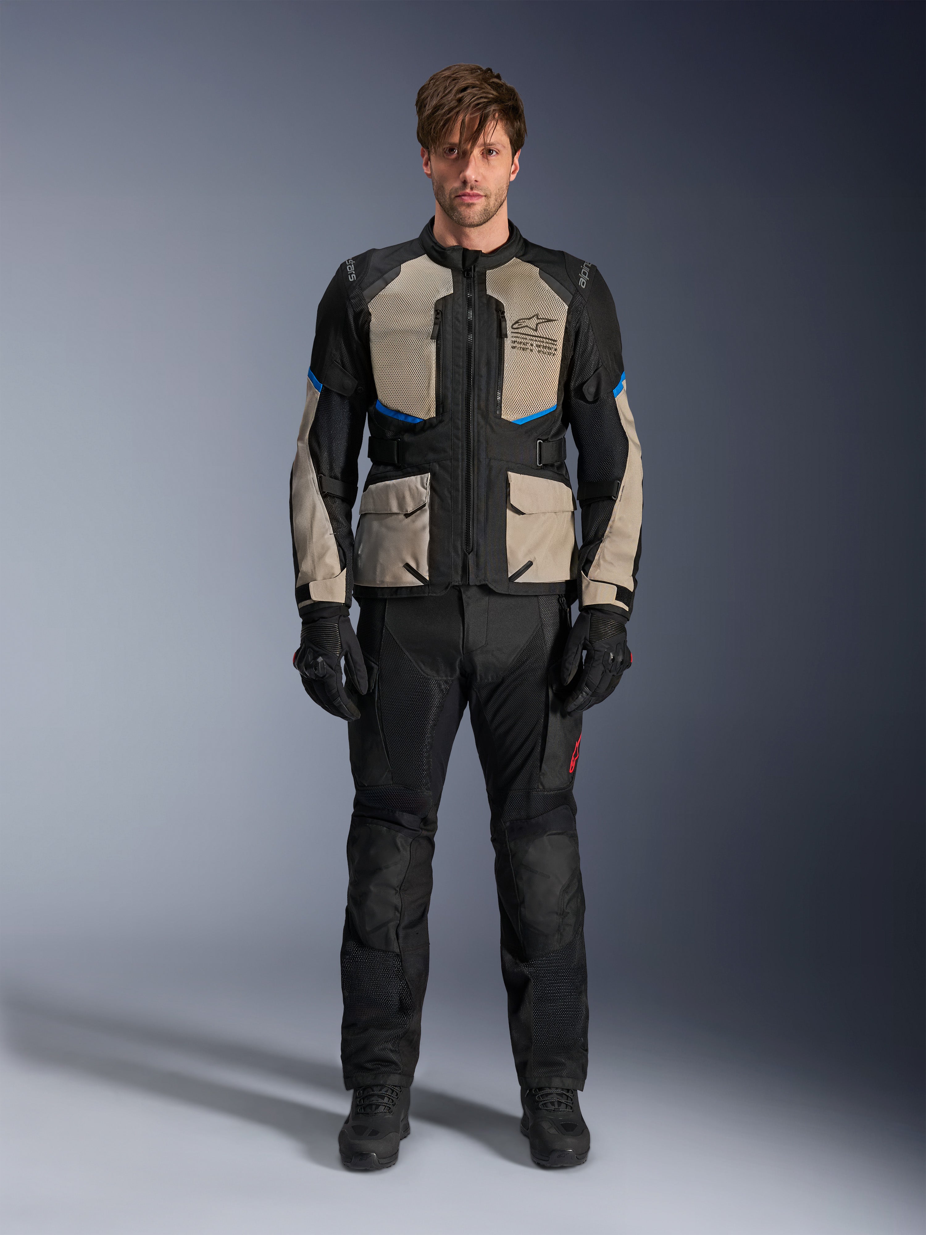 Model draagt Alpinestars Andes Air Drystar® jas, motor touringjas, Black Laurel Gray en Bright Blue, gecombineerd met bijpassende zwarte rijbroek en handschoenen, voorzien van uitgebreide mesh ventilatiepanelen op de borst en mouwen voor adventure touring in warm weer.