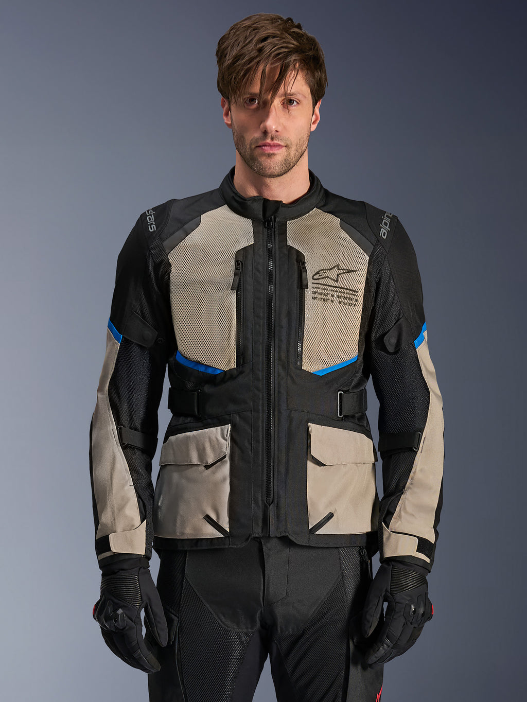 Alpinestars Andes Air Drystar motorjas, adventure touringjas, zwart, Laurel grijs en helderblauw, gedragen door een model met zwarte handschoenen, voorzien van grote mesh borstpanelen voor luchtstroom, cargo-zakken aan de voorzijde en blauwe accenten, beschermende motorkleding