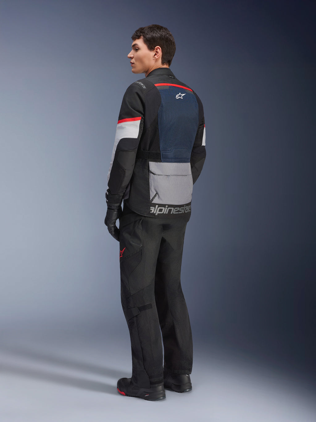 Mannelijk model van achteren en opzij gezien, dragend de Alpinestars Andes Air Drystar® Jas in Deep Blue Black Ice Gray, gecombineerd met een zwarte motorbroek en handschoenen, voorzien van een blauw mesh achterpaneel, grijze opbergzak op de onderrug en rode accenten op de mouwen, professionele adventure touring uitrusting.