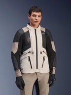 Alpinestars AMT-10LAB Drystar® XF jas, White Sand Aluminum Camo, adventure motorjas met zwarte MATRYX® mesh panelen en lederen verstevigingen, gedragen door een model met zwarte handschoenen en bijpassende zandkleurige broek, studioportret