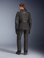Vrouwelijke motorrijder van achteren gezien in de Alpinestars Stella Andes V4 Drystar® jas, zwart, gecombineerd met een bijpassende motorbroek, wit Astar-logo op de rug, verlengde achterzoom, complete adventure-touring motoroutfit inclusief zwarte handschoenen en laarzen