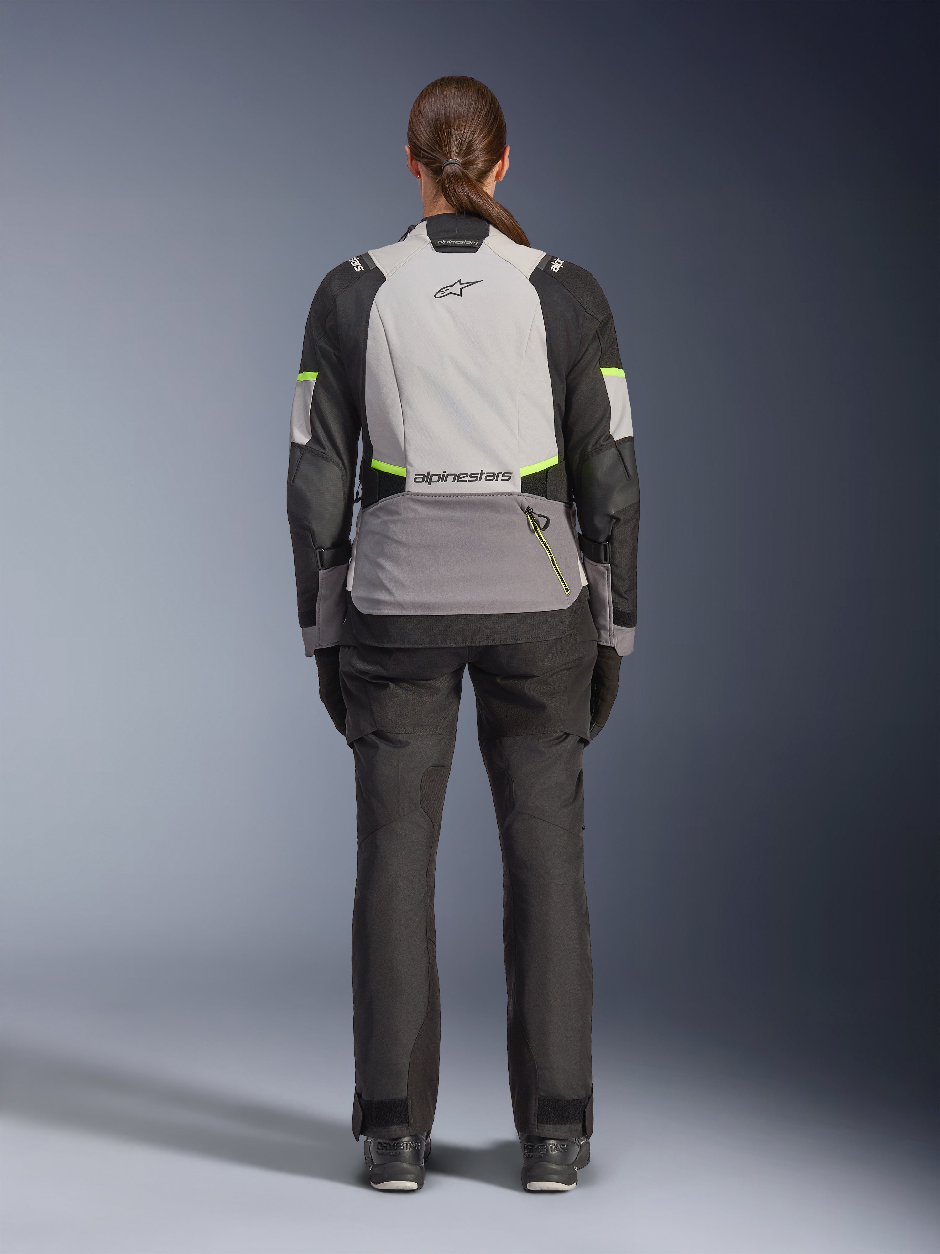 Vrouwelijke motorrijder vanaf de achterkant in de Alpinestars Stella Andes V4 Drystar® Jas, Ice Gray, Dark Gray en zwart met Yellow Fluo accenten, voorzien van een grote opbergzak op de rug en Alpinestars-logo, gecombineerd met een bijpassende zwarte adventure-broek, all-weather motoruitrusting voor touring