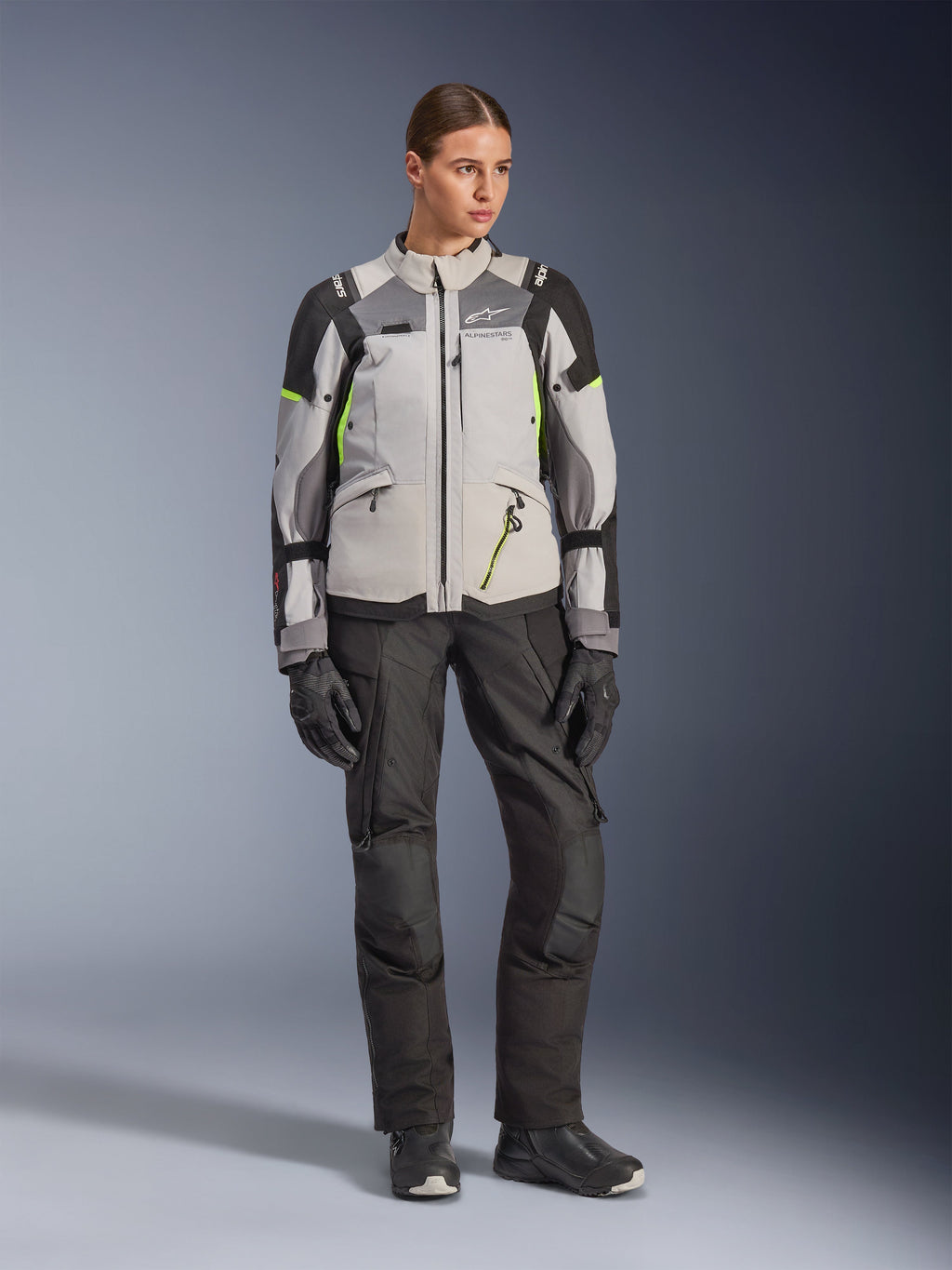 Vrouwelijk model draagt Alpinestars Stella Andes V4 Drystar® Jas in Ice Gray, Dark Gray en zwart met Yellow Fluo accenten, gecombineerd met een zwarte motorbroek en handschoenen, volledige studio-opname van adventure touring-uitrusting.
