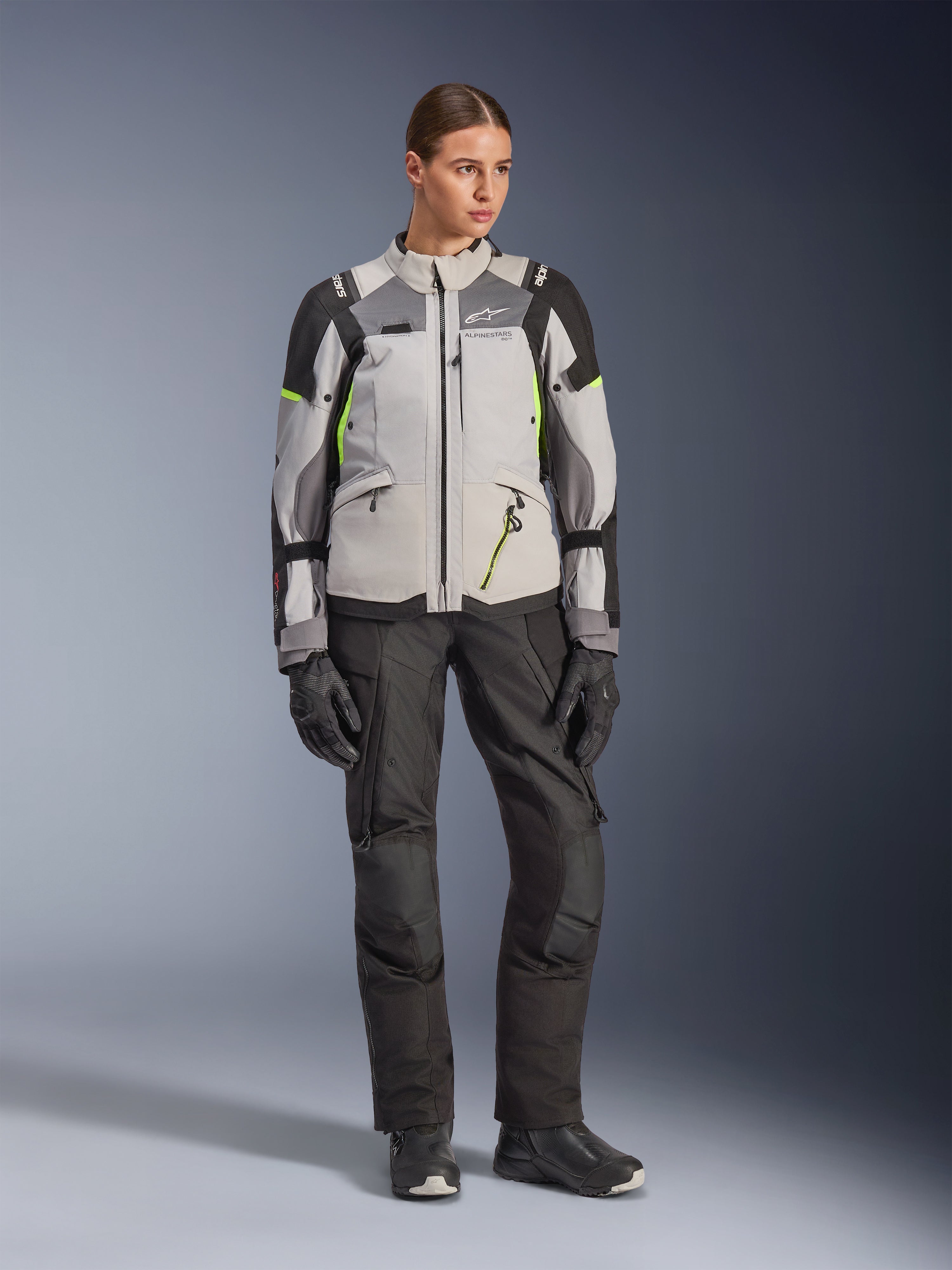 Vrouwelijk model draagt Alpinestars Stella Andes V4 Drystar® Jas in Ice Gray, Dark Gray en zwart met Yellow Fluo accenten, gecombineerd met een zwarte motorbroek en handschoenen, volledige studio-opname van adventure touring-uitrusting.