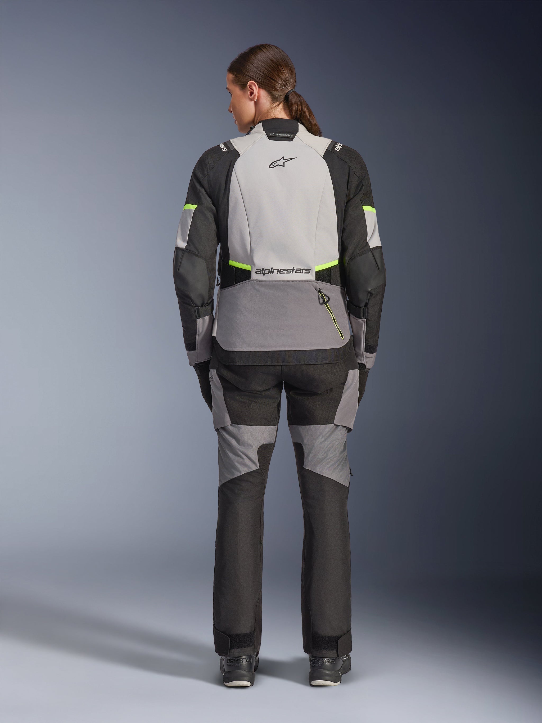 Female rider from the back wearing Alpinestars Stella Andes V4 Drystar® Jacket and matching pants, Ice Gray and Dark Gray with Black and Yellow Fluo accents, adventure-touring motorcycle gear with durable textile construction and reflective branding