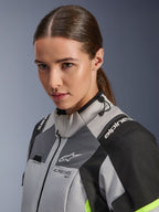 Alpinestars Stella Andes V4 Drystar® Jas, Ice Gray en Dark Gray met zwarte en geel fluo accenten, close-up van een vrouwelijk model dat de waterdichte motorjas draagt, Alpinestars-branding op de borst en schouders, verstelbare kraag met trekkoord