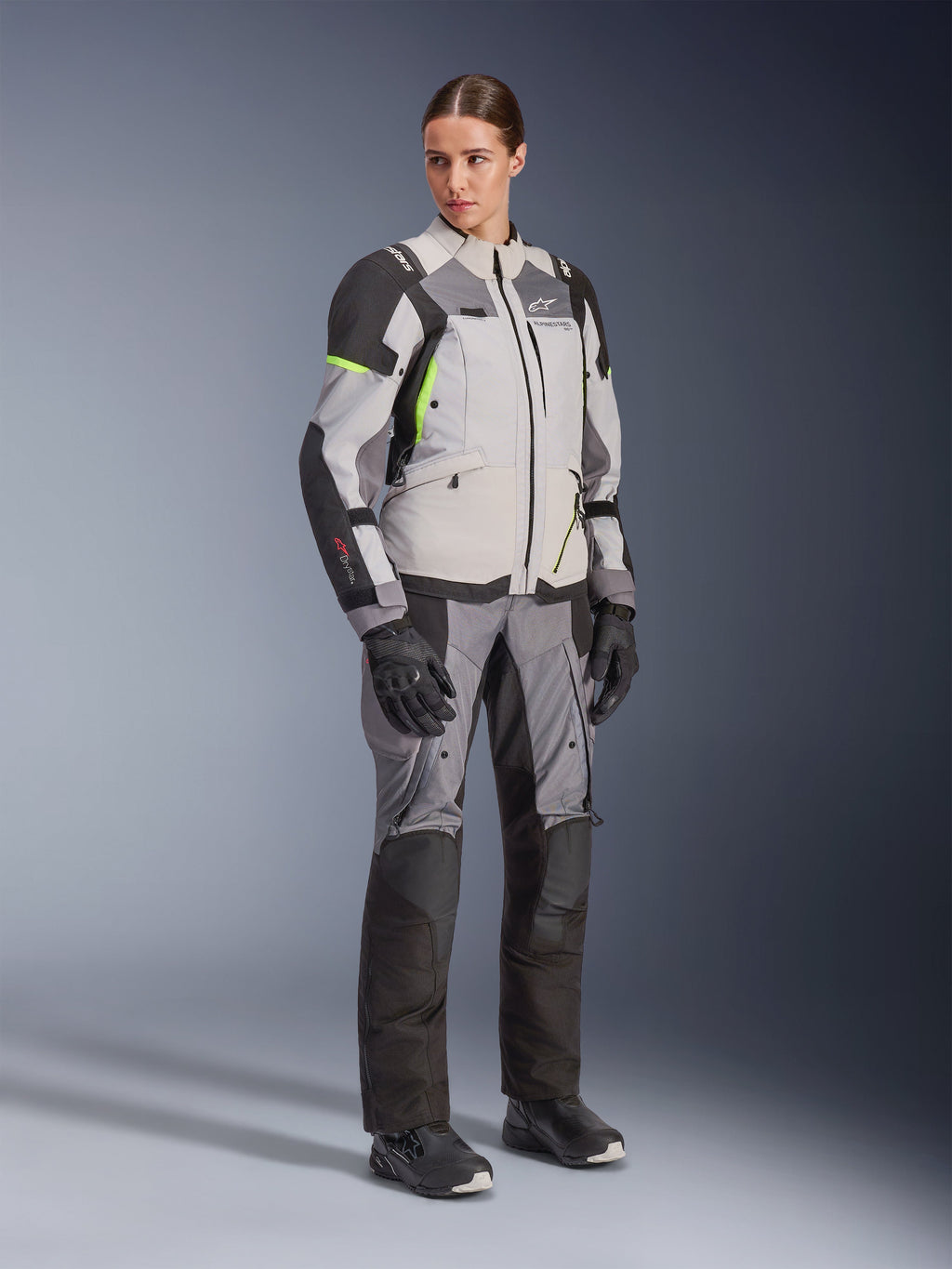 Vrouw draagt Alpinestars Stella Andes V4 Drystar® jas en bijpassende broek, Ice Gray en Dark Gray met fluorgele accenten, complete adventure touring kledingset met zwarte handschoenen en laarzen, volledige studiofoto