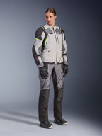 Vrouw draagt Alpinestars Stella Andes V4 Drystar® jas en bijpassende broek, Ice Gray en Dark Gray met fluorgele accenten, complete adventure touring kledingset met zwarte handschoenen en laarzen, volledige studiofoto