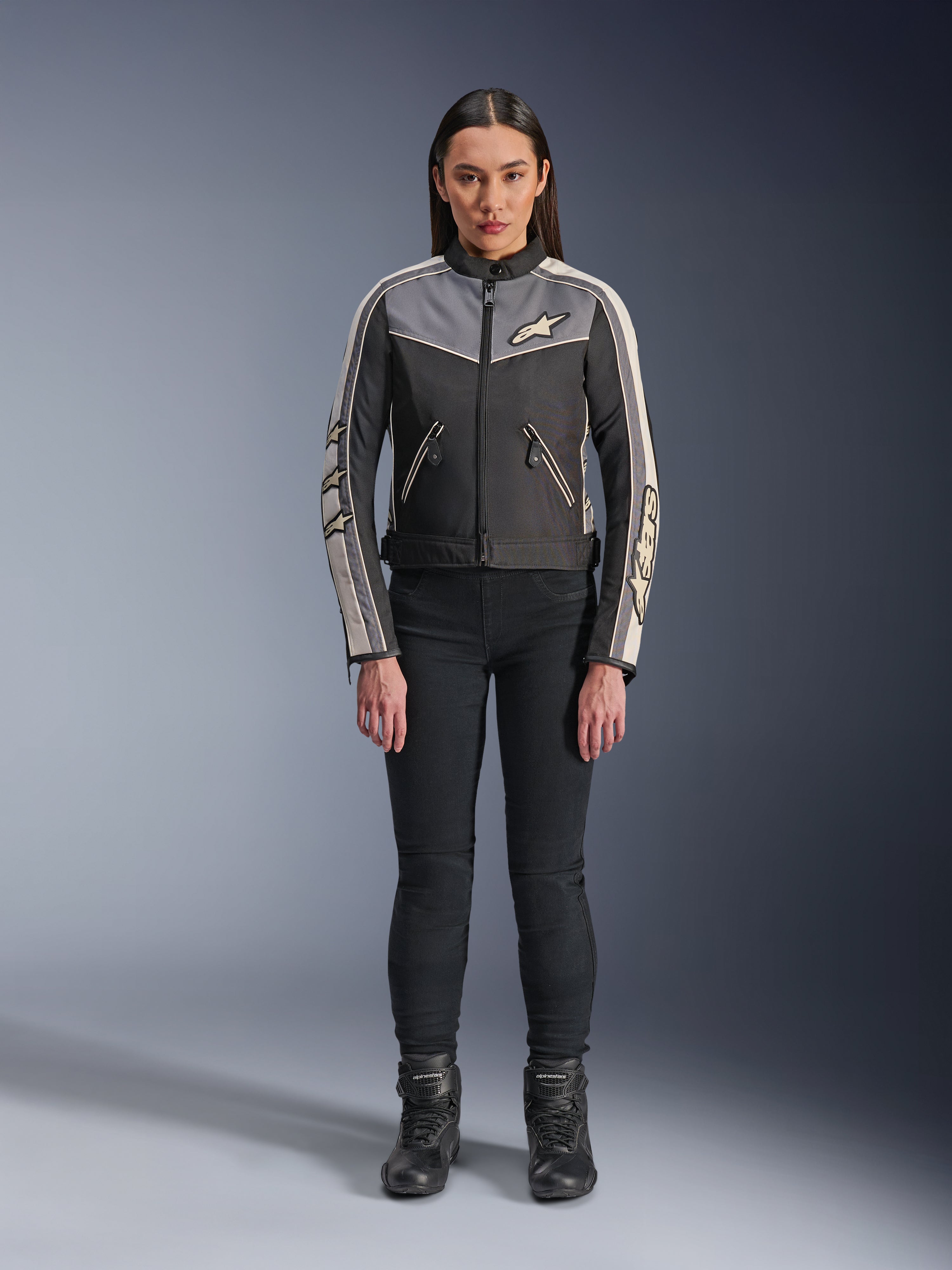 Vrouw in Alpinestars Stella T-Dyno WR-jas, zwart, donkergrijs en ecru, vooraanzicht, textiele motorjas met een op vintage geïnspireerd ontwerp, voorzien van merklogo's op de borst en mouwen, gedragen met een zwarte broek en laarzen