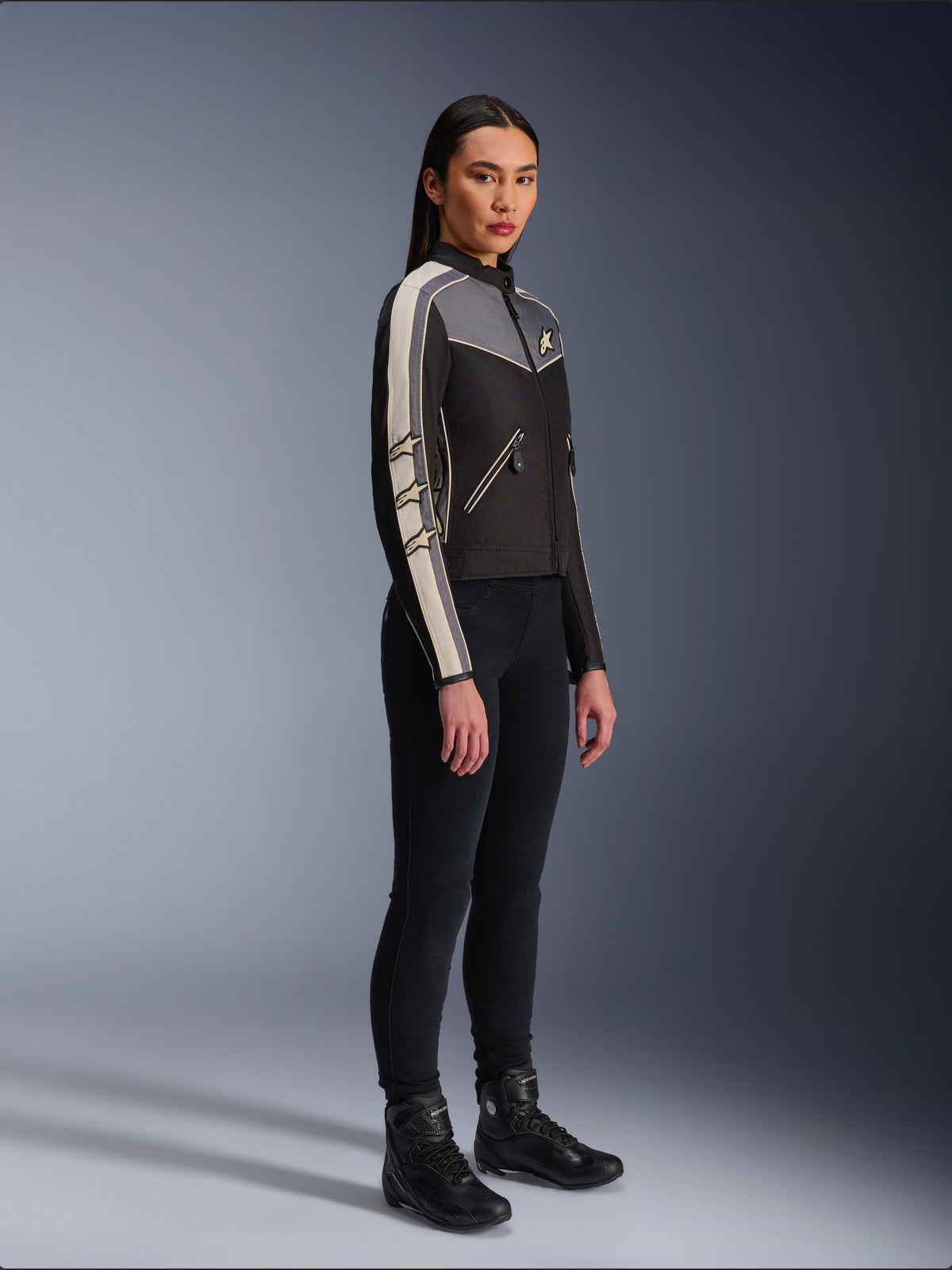 Alpinestars Stella T-Dyno WR Jas, textiele motorjas voor dames, zwart, donkergrijs en ecru, met een op vintage geïnspireerd ontwerp met crèmekleurige strepen op de mouwen en ster-logo's, gedragen met een zwarte broek en motorlaarzen, op erfgoed geïnspireerde urban motorkleding