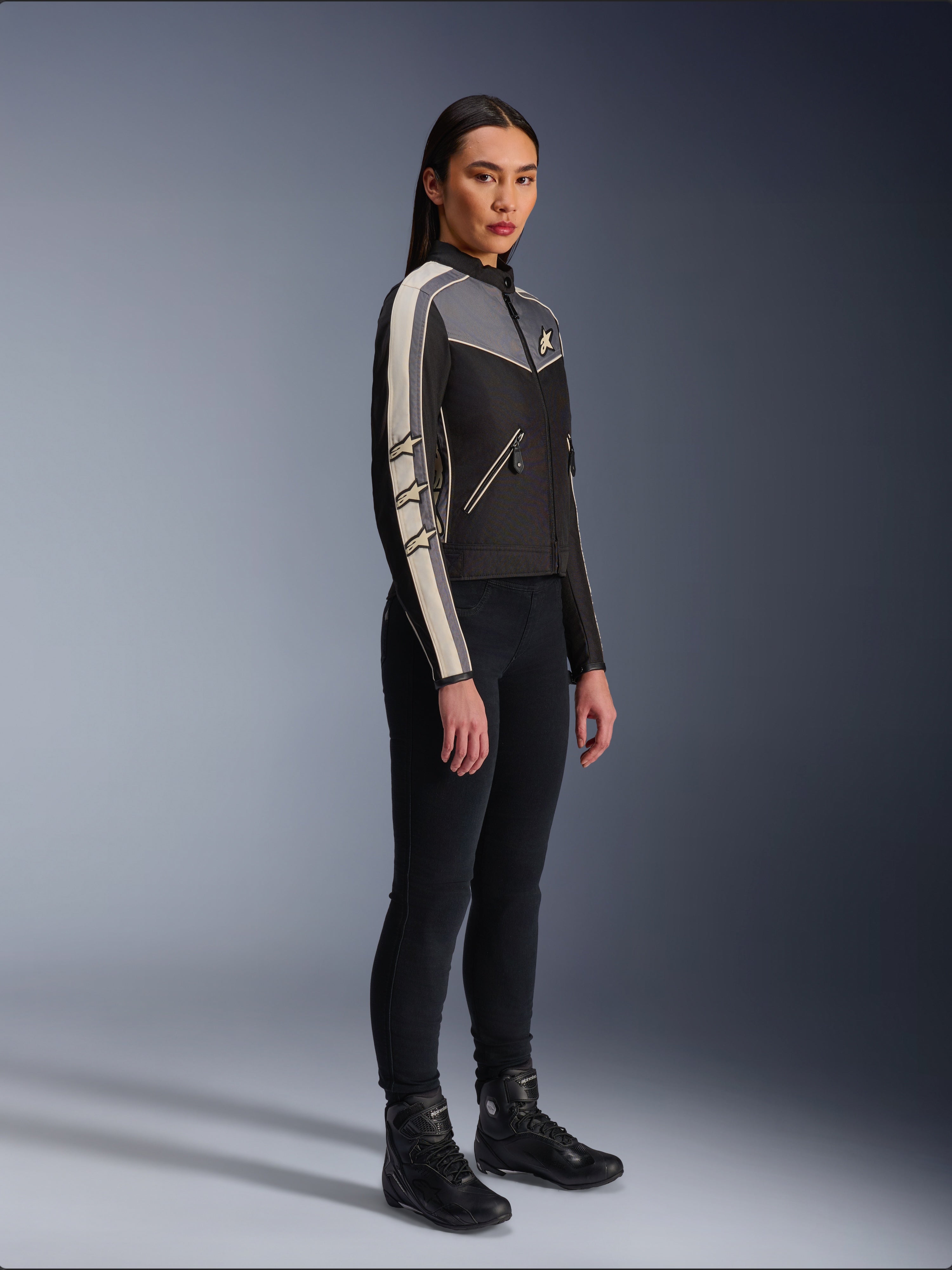 Alpinestars Stella T-Dyno WR Jas, textiele motorjas voor dames, zwart, donkergrijs en ecru, met een op vintage geïnspireerd ontwerp met crèmekleurige strepen op de mouwen en ster-logo's, gedragen met een zwarte broek en motorlaarzen, op erfgoed geïnspireerde urban motorkleding