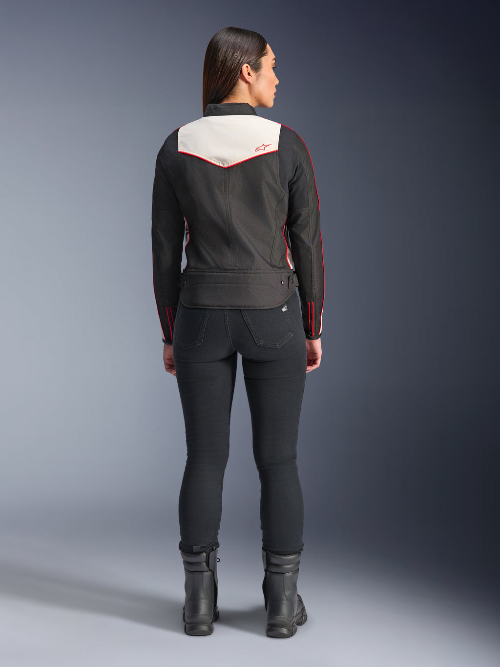 Achteraanzicht van een vrouwelijk model in de Alpinestars Stella T-Dyno WR-jas in Black Ecru Bright Red, voorzien van een opvallend crème rugpaneel en rode logo-accenten, gecombineerd met een zwarte broek en laarzen, een vintage-geïnspireerde textiele motorjas.