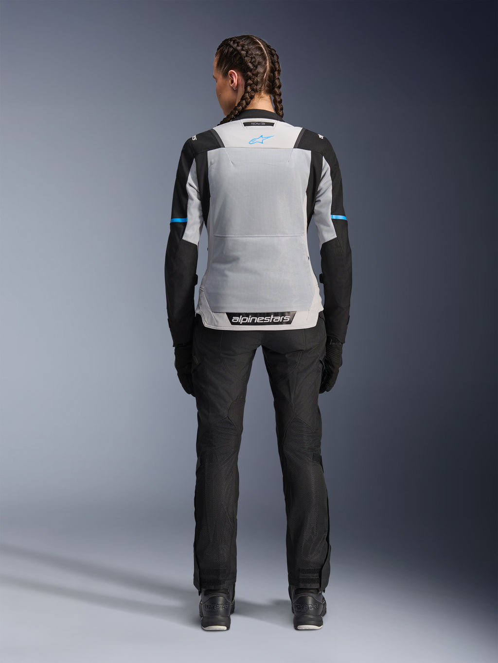 Alpinestars Stella ST-2 Air Jacket, Ice Gray Black, achteraanzicht op vrouwelijk model, voorzien van een groot lichtgrijs mesh rugpaneel, zwarte mouwen met blauwe streepaccenten, Alpinestars-branding op de onderrug, gecombineerd met zwarte technische motorbroek en laarzen, zomerse motoruitrusting
