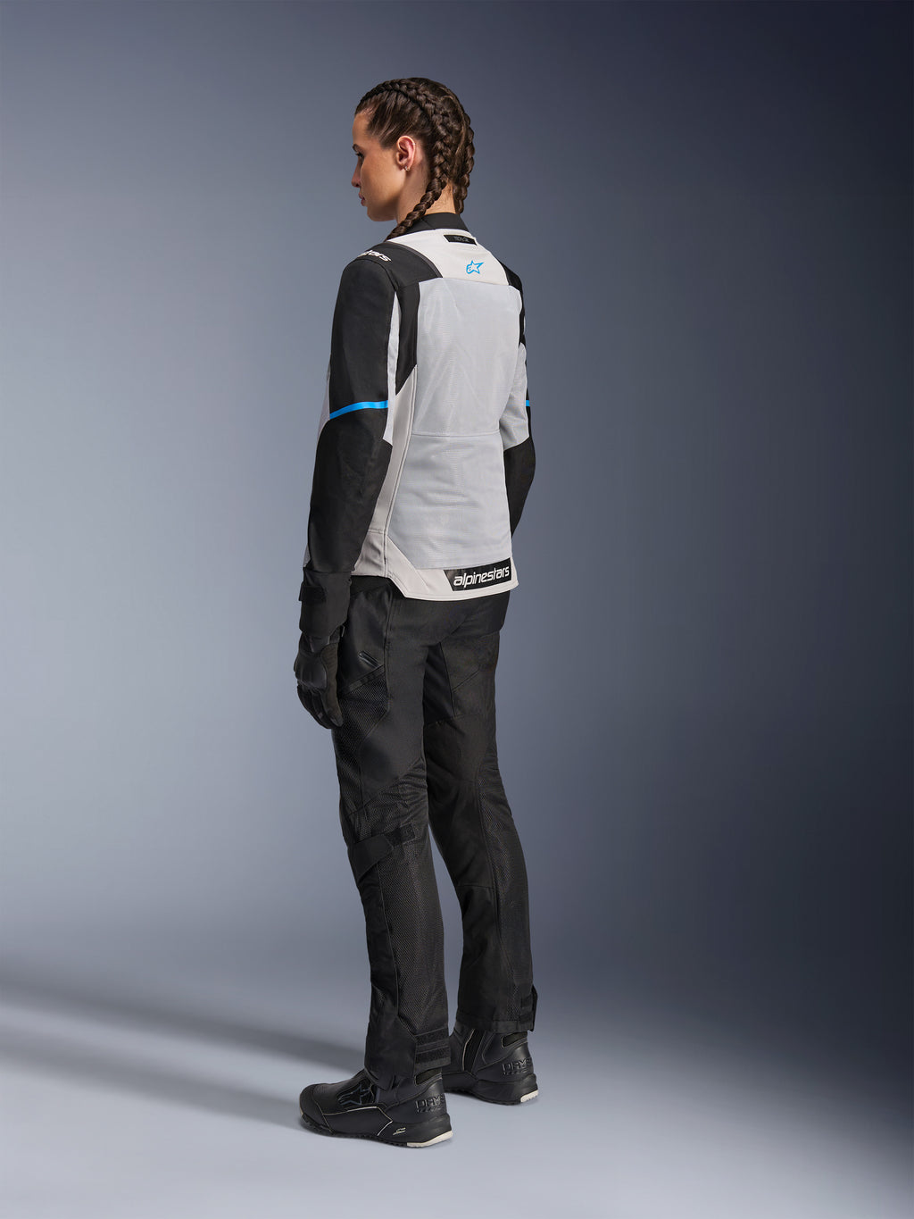 Vrouwelijke motorrijder draagt het Alpinestars Stella ST-2 Air Jacket in Ice Gray en zwart, driekwart achteraanzicht met focus op het grote lichtgrijze mesh rugpaneel, zwarte mouwen met een blauwe accentstreep en een blauw Alpinestars-logo, gecombineerd met een zwarte motorbroek, handschoenen en laarzen voor een complete adventure touring-outfit
