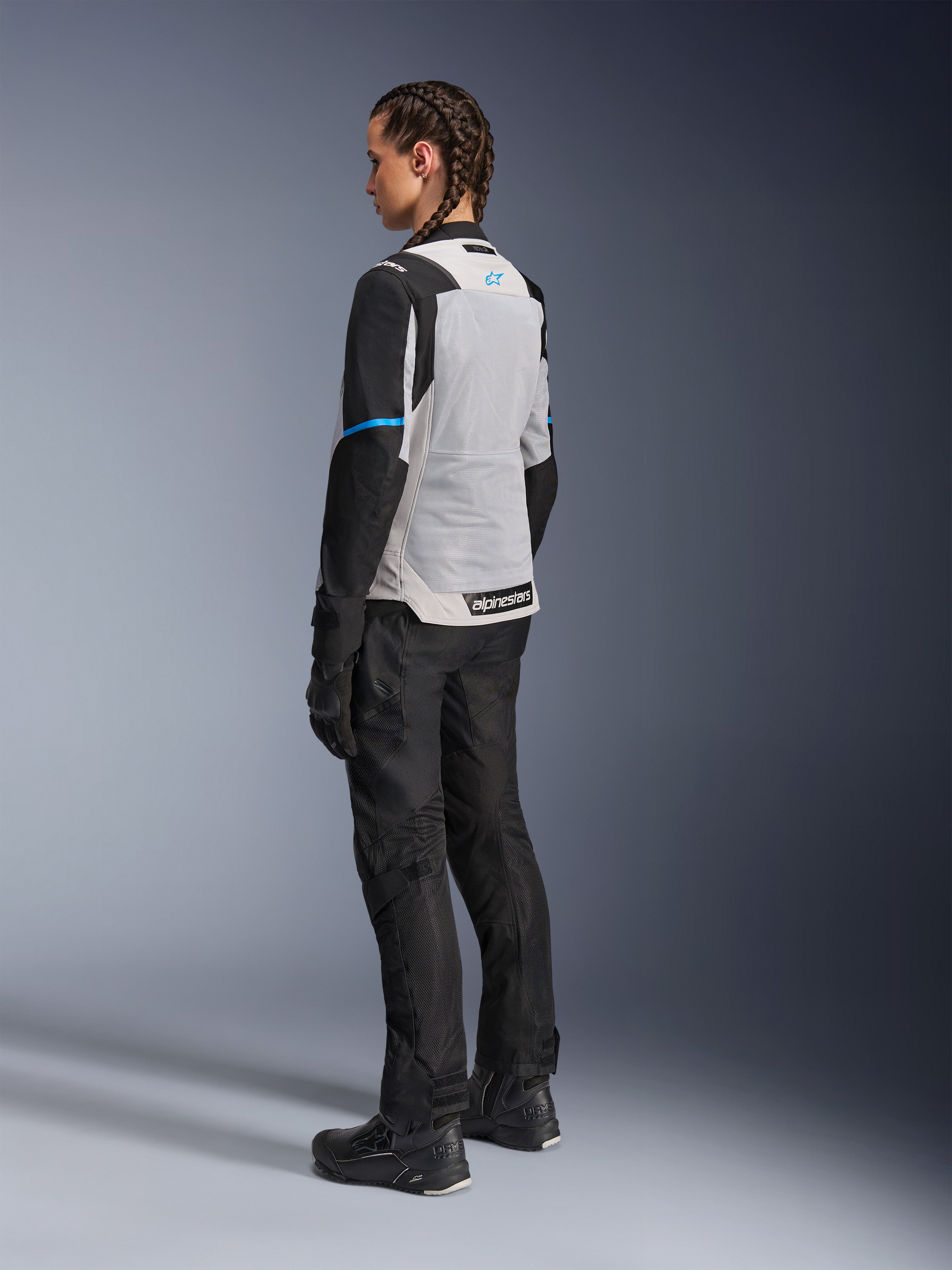 Vrouwelijke motorrijder draagt het Alpinestars Stella ST-2 Air Jacket in Ice Gray en zwart, driekwart achteraanzicht met focus op het grote lichtgrijze mesh rugpaneel, zwarte mouwen met een blauwe accentstreep en een blauw Alpinestars-logo, gecombineerd met een zwarte motorbroek, handschoenen en laarzen voor een complete adventure touring-outfit