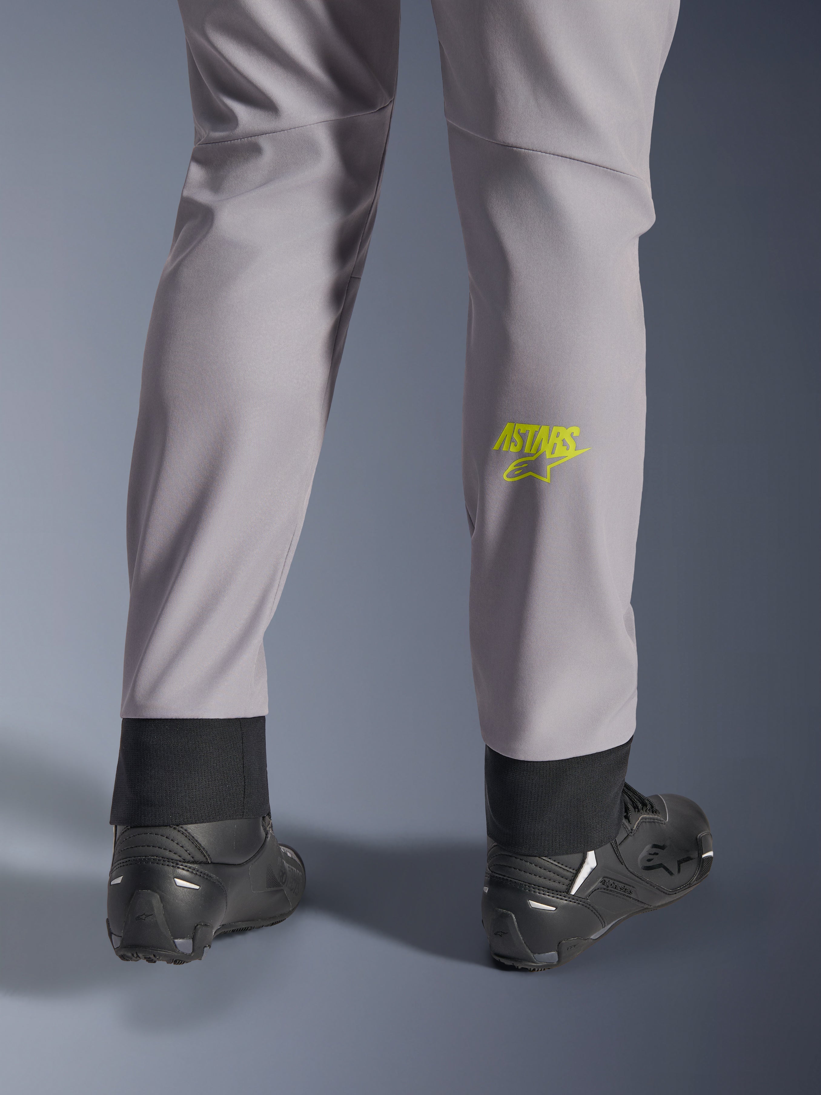 Alpinestars Aeroshell Pants, Mid Gray with Yellow Fluo Astars logo on the calf, shown from the back wearing black motorcycle shoes, featuring an elastic lower hem and a modern tapered silhouette