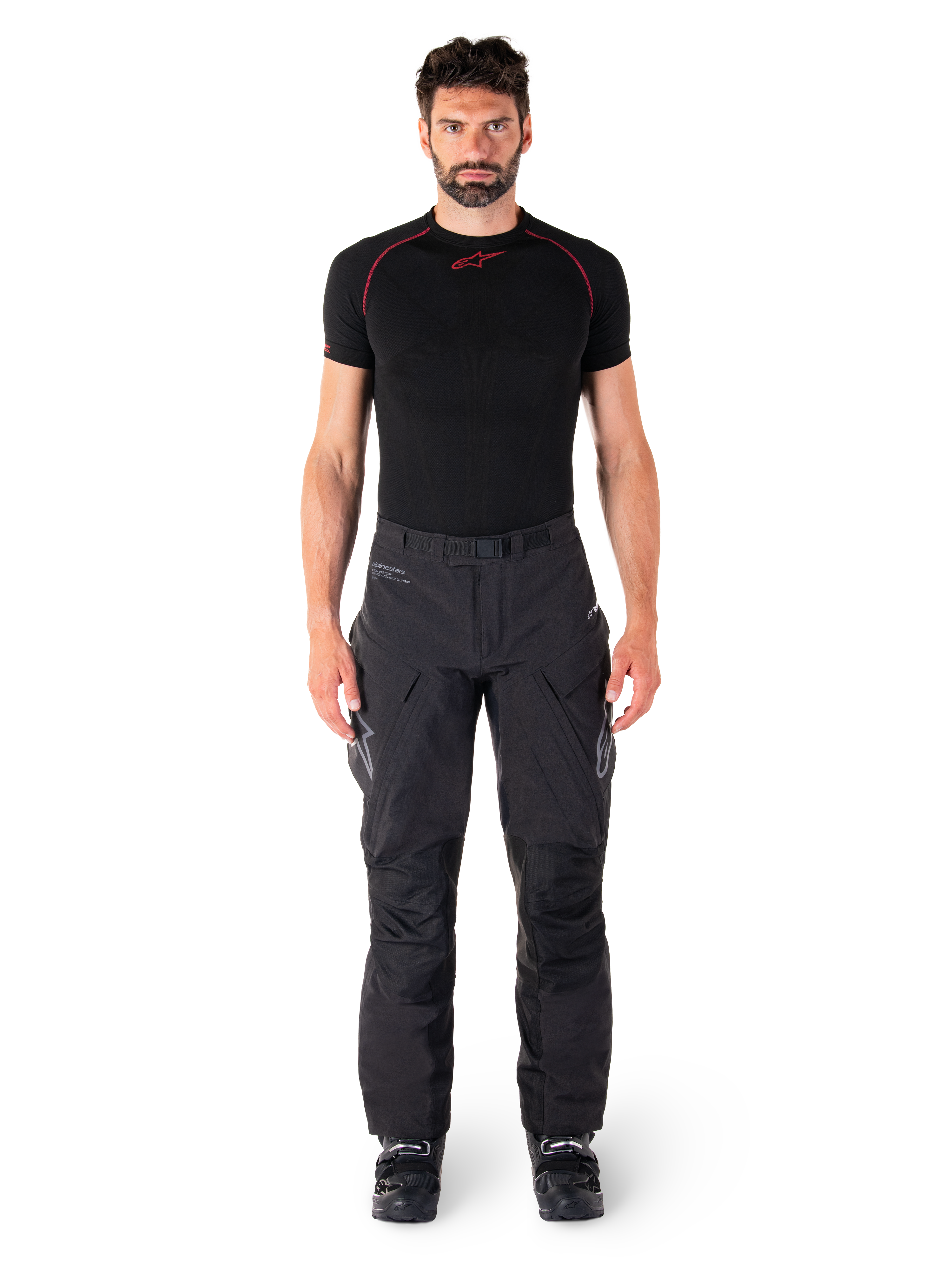 Alpinestars Hyde XT Drystar® Broek, adventure touring motorbroek, zwart, gedragen door een model met een zwarte basislaag met korte mouwen en zwarte laarzen, voorzien van technisch stretchmateriaal, versterkte kniepanelen en ergonomische cargozakken.