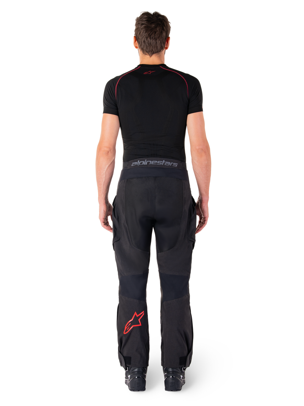 Alpinestars Hyde Xt Drystar® broek, adventure touring motorbroek, zwart en felrood, achteraanzicht van een rijder die de broek en een zwarte basislaag draagt, met een rood sterlogo op het linkerbeen en grijze merknaam op de tailleband.