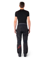 Alpinestars Hyde Xt Drystar® broek, adventure touring motorbroek, zwart en felrood, achteraanzicht van een rijder die de broek en een zwarte basislaag draagt, met een rood sterlogo op het linkerbeen en grijze merknaam op de tailleband.