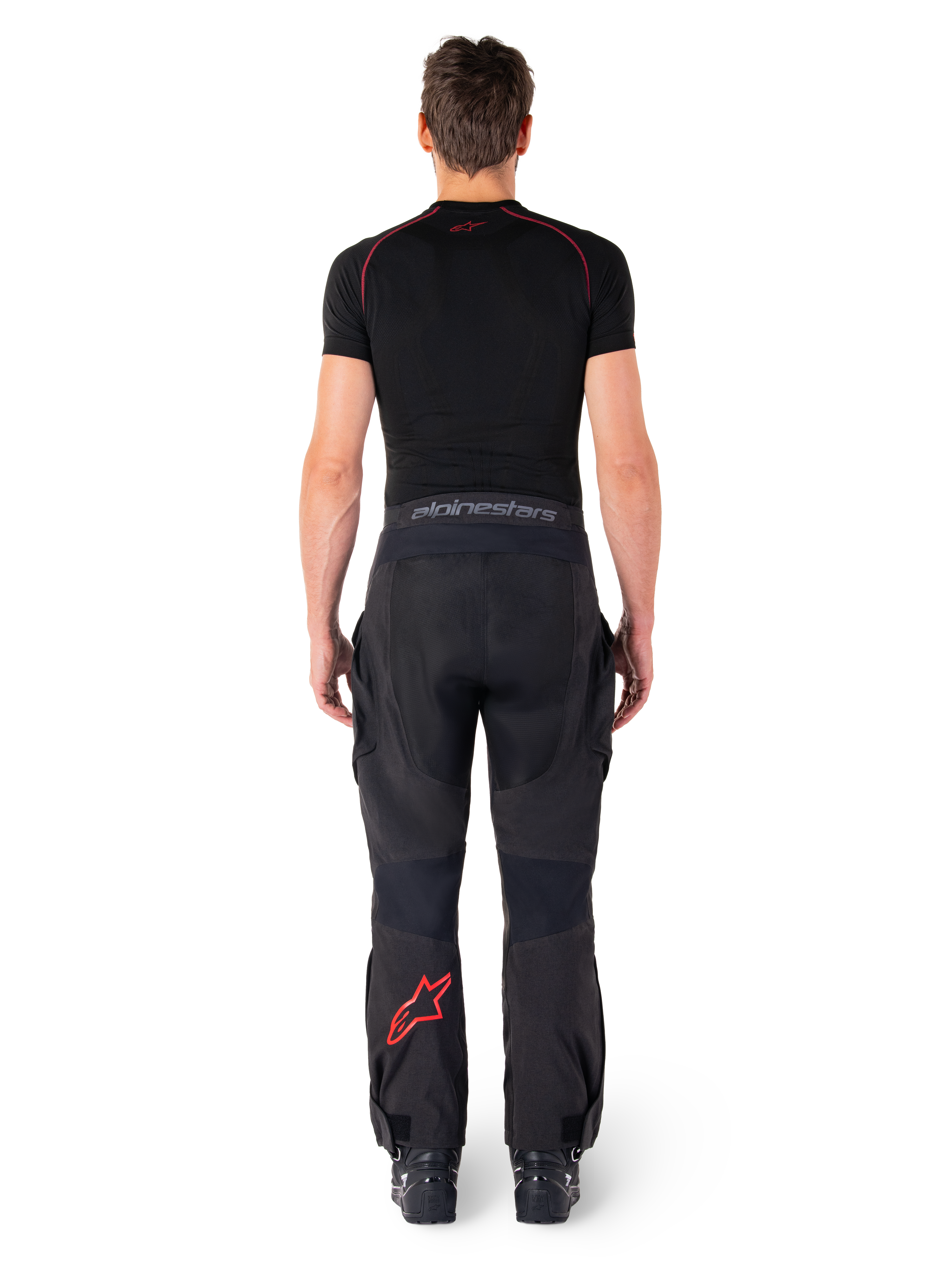 Alpinestars Hyde Xt Drystar® broek, adventure touring motorbroek, zwart en felrood, achteraanzicht van een rijder die de broek en een zwarte basislaag draagt, met een rood sterlogo op het linkerbeen en grijze merknaam op de tailleband.
