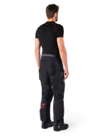 Alpinestars Hyde XT Drystar® broek, adventure touring broek, zwart en felrood, achteraanzicht van een rijder die de broek draagt met een zwarte basislaag en zwarte laarzen, met een duurzame textielconstructie, een rood sterlogo op het onderbeen en grijze Alpinestars-branding op de tailleband