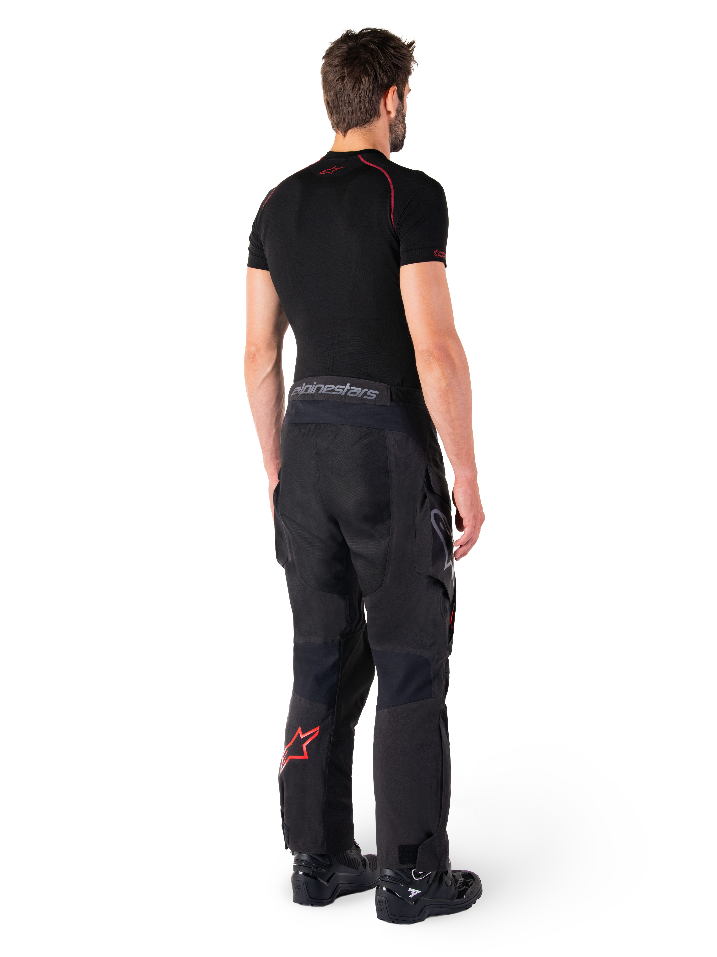 Alpinestars Hyde XT Drystar® broek, adventure touring broek, zwart en felrood, achteraanzicht van een rijder die de broek draagt met een zwarte basislaag en zwarte laarzen, met een duurzame textielconstructie, een rood sterlogo op het onderbeen en grijze Alpinestars-branding op de tailleband
