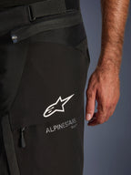 Close-up detail van de Alpinestars Andes V4 Drystar® broek in het zwart, met het witte Alpinestars-logo en woordmerk op de flap van een cargozak, met zichtbare tailleverstelband en verbindingsrits, ontworpen voor adventure motorrijden.
