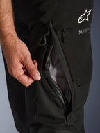 Close-up van de Alpinestars Andes V4 Drystar® broek in het zwart, met een hand die de directe ventilatieopening op de dij opent met mesh-voering, wat de duurzame textielconstructie en Alpinestars-branding benadrukt.