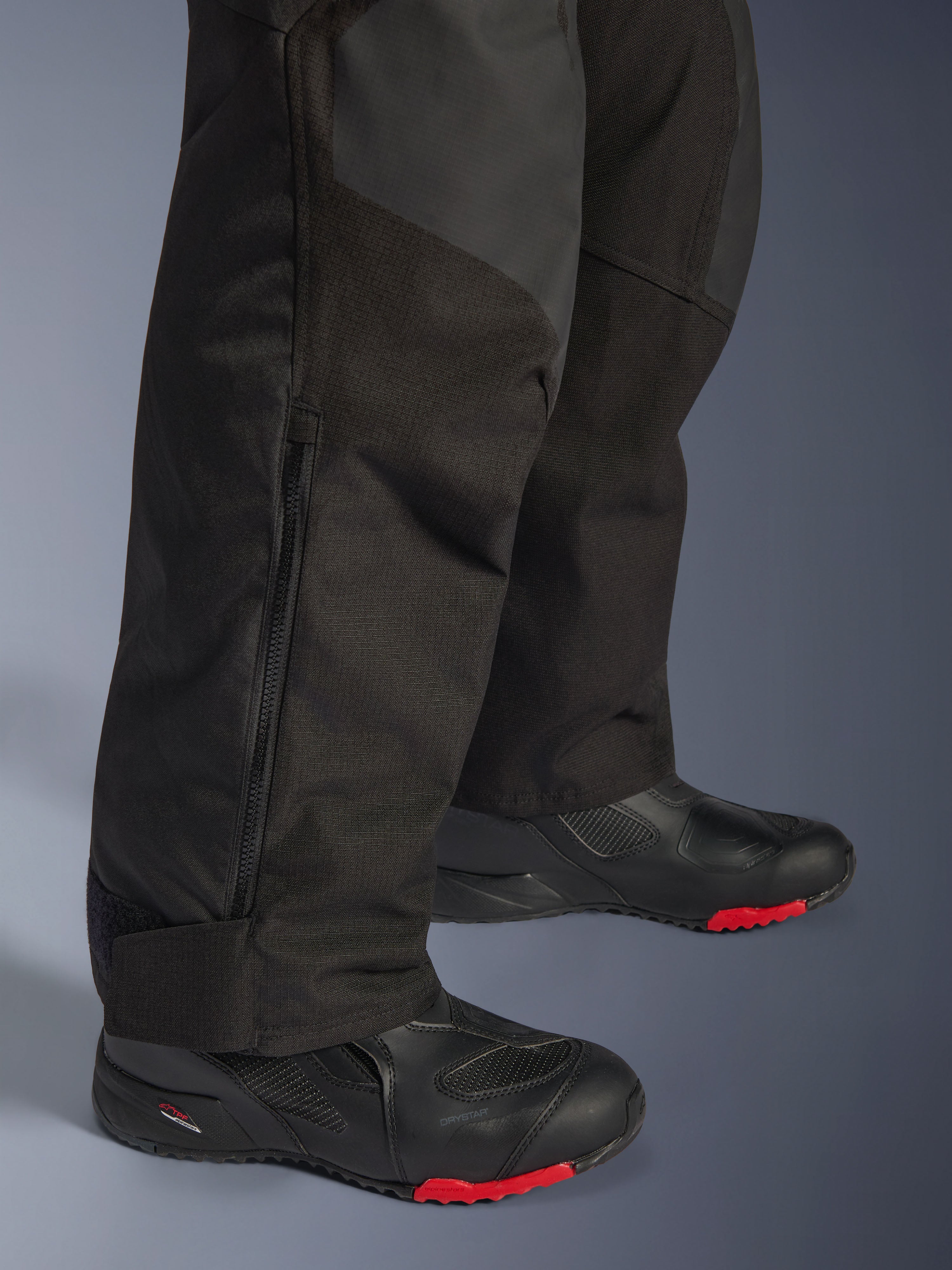 Alpinestars Andes V4 Drystar® Broek - Lang, zwarte adventure motorbroek, close-up van het onderbeen met verstelbare ritssluiting bij de enkel en duurzame textielconstructie, gedragen met zwarte motorlaarzen met rode accenten