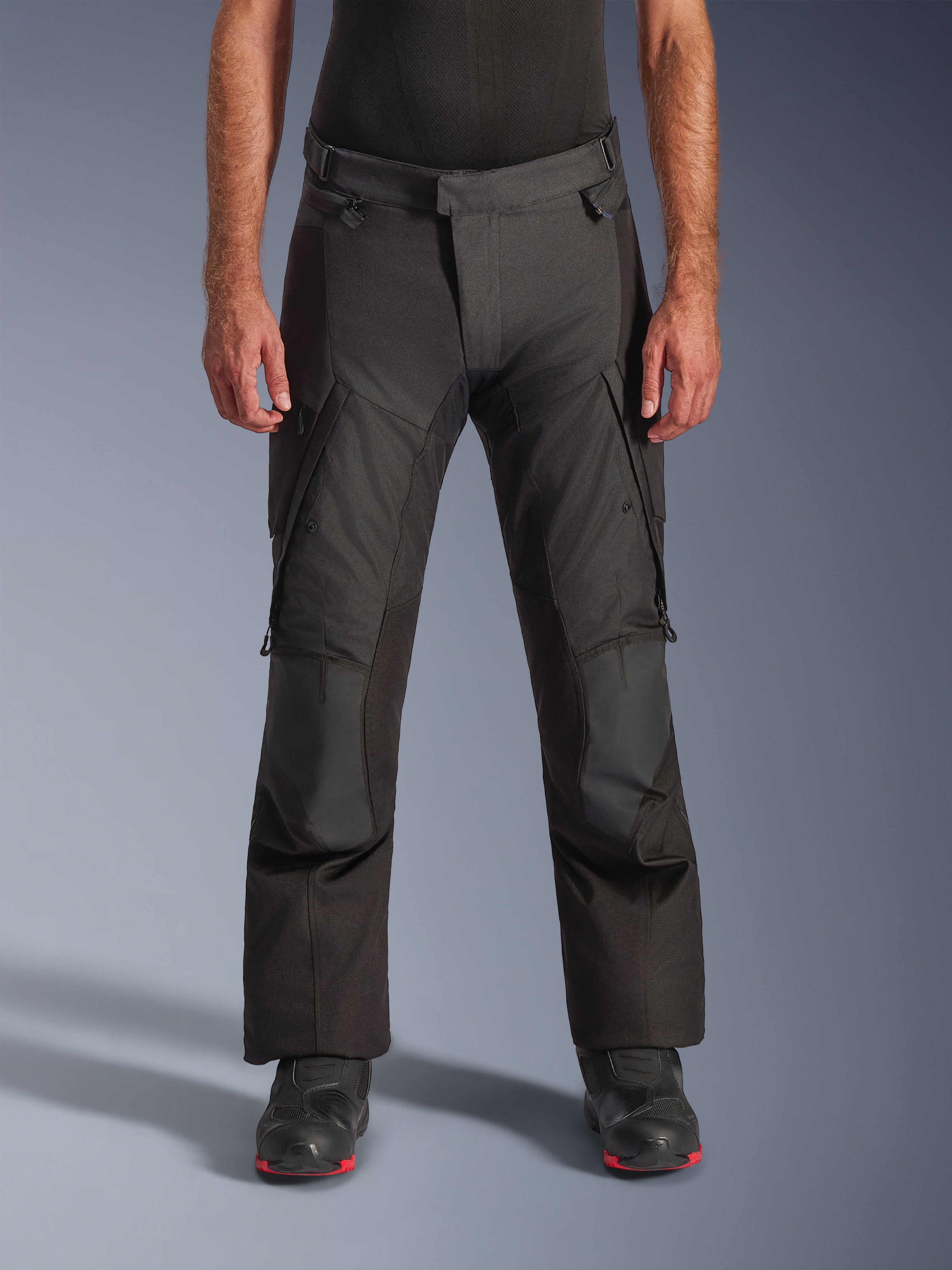 Alpinestars Andes V4 Drystar motorbroek, adventure touring motorbroek, zwart met grijze panelen, voorzien van verstevigde kniestukken en cargozakken op de dijen, gedragen met zwarte motorlaarzen, waterdichte textielconstructie voor alle weersomstandigheden