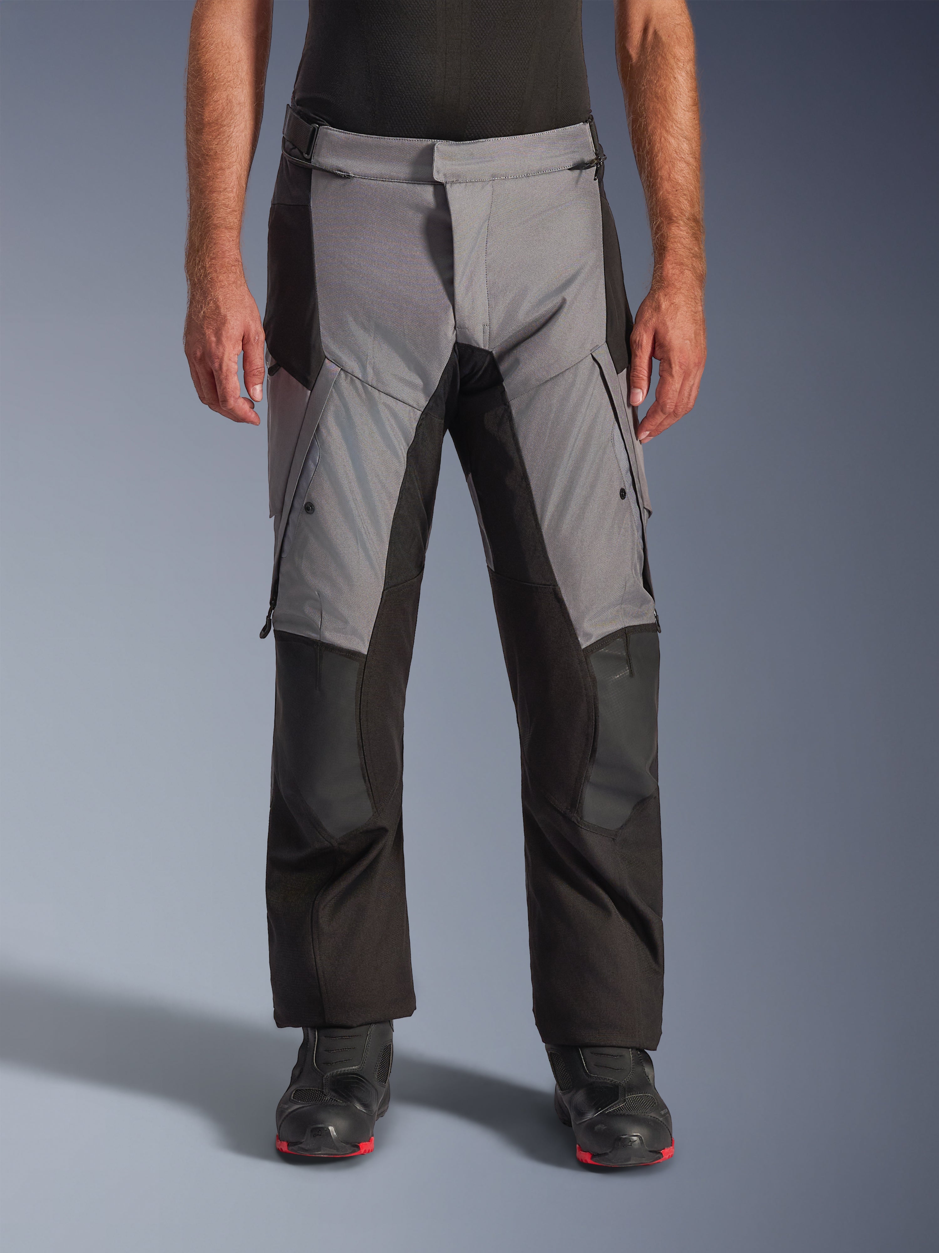 Alpinestars Andes V4 Drystar® broek, adventure touring motorbroek, grijs en zwart, vooraanzicht gedragen door een model met zwarte motorlaarzen, voorzien van versterkte kniepanelen en cargozakken op de dijen, waterdichte ademende textielconstructie voor alle weersomstandigheden