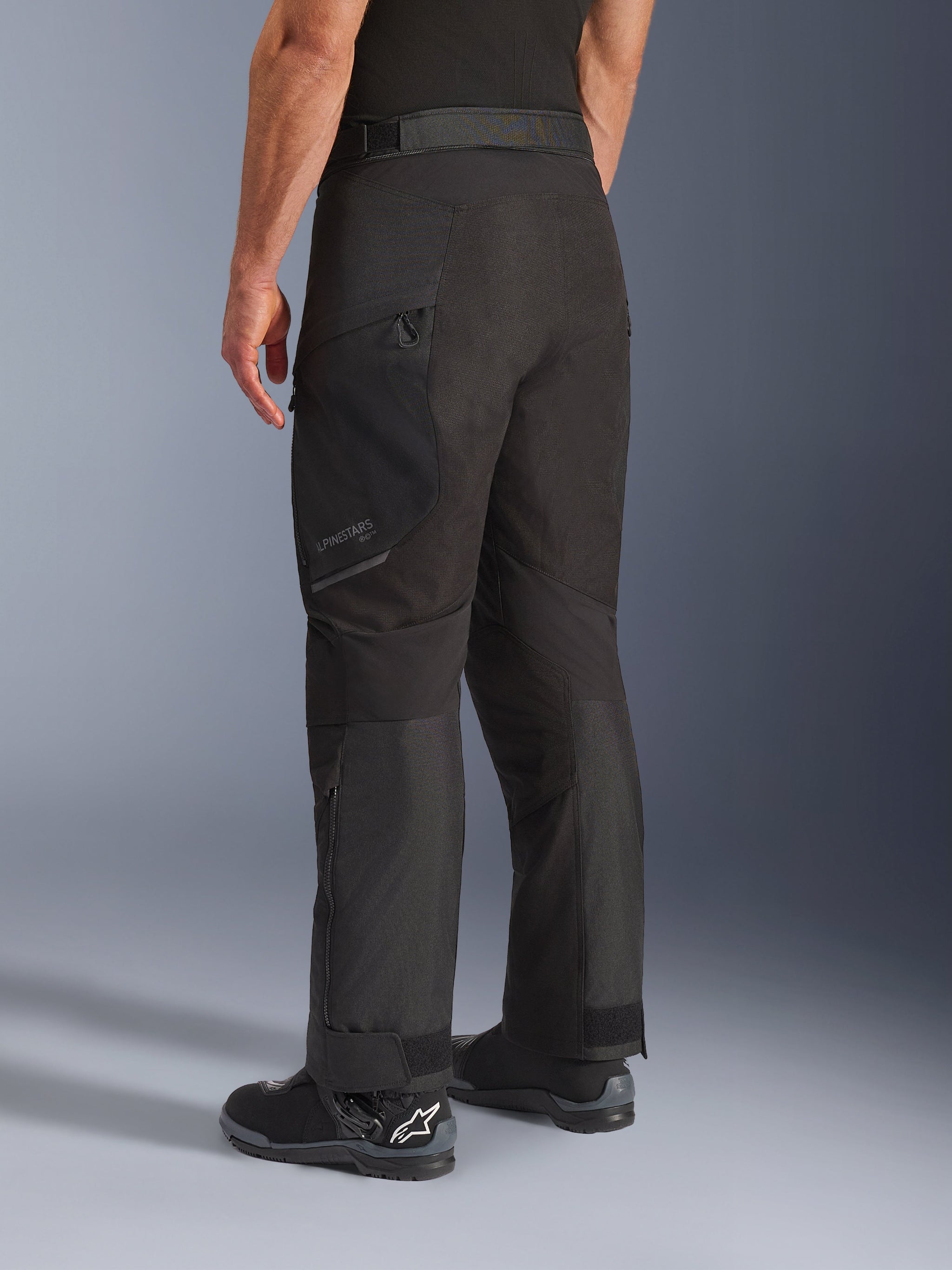 Alpinestars Cusco Drystar® broek, motorbroek, zwart, driekwart achteraanzicht met dijbeenzak met ritssluiting en voorgevormde panelen, gedragen met zwarte motorlaarzen, duurzaam adventure touring-ontwerp met subtiele branding