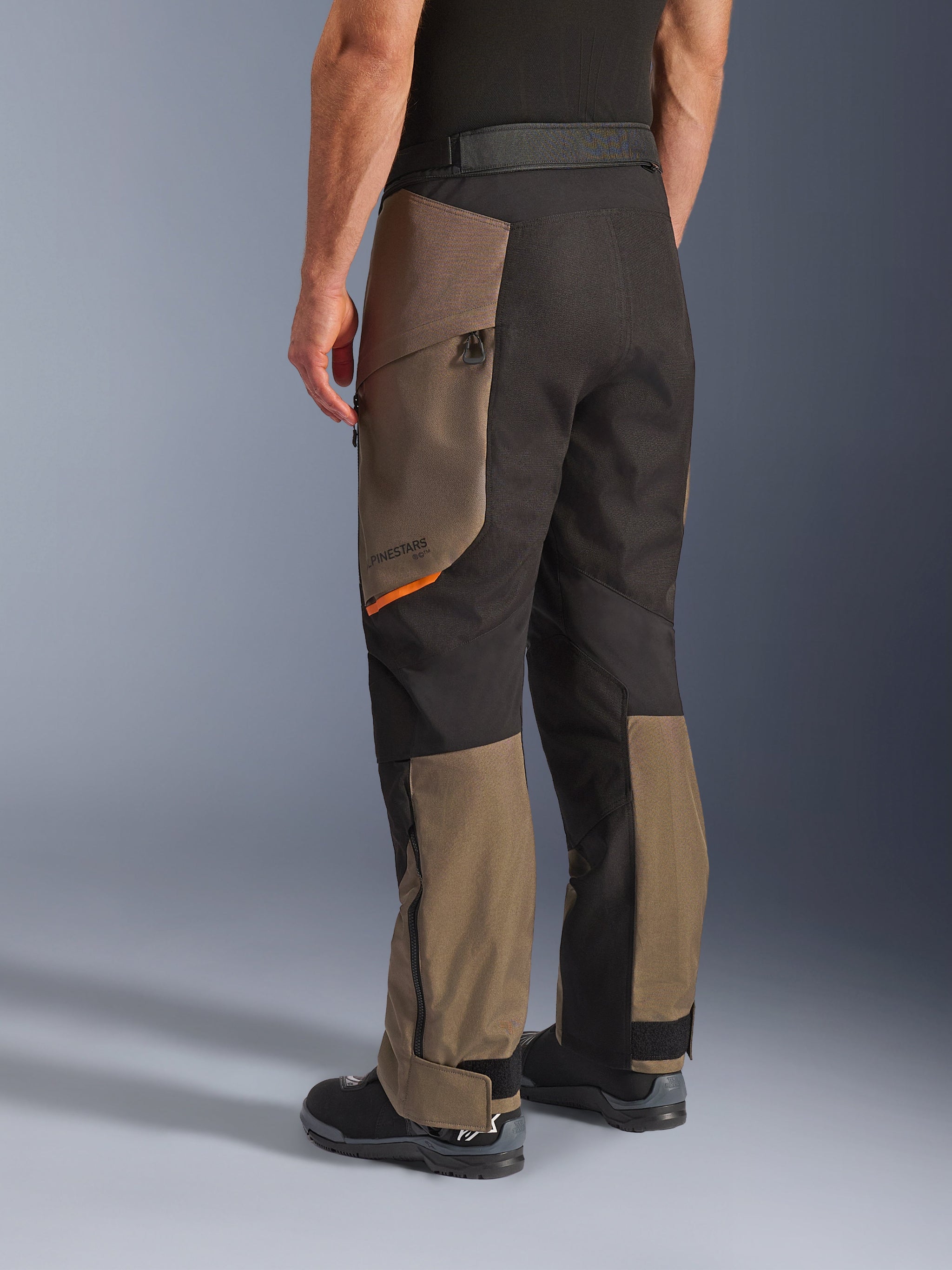 Alpinestars Cusco Drystar® broek, adventure motorbroek, Walnut Black met klein oranje accent, driekwart achteraanzicht toont de textielconstructie en walnut panelen, gedragen met zwarte motorlaarzen