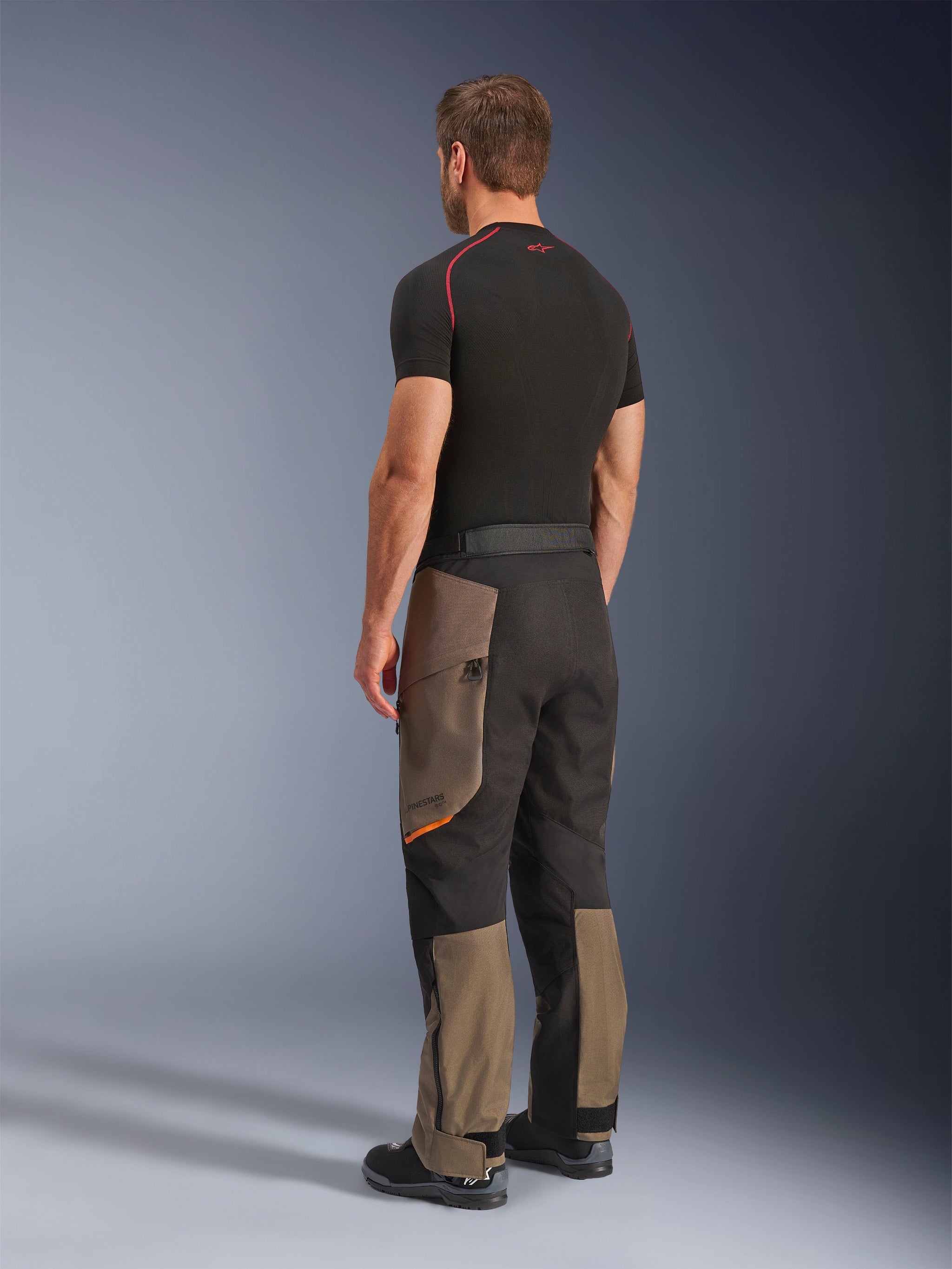 Alpinestars Cusco Drystar® broek, adventure motorbroek, Walnut Black, driekwart achteraanzicht gedragen met een zwarte basislaag en laarzen, voorzien van versterkte textielpanelen, cargozakken met rits op de bovenbenen en een ergonomische touring-pasvorm.