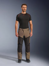 Alpinestars Cusco Drystar® broek, Walnut Black, adventure touring motorbroek, gedragen door een mannelijk model met een zwarte basislaag en zwarte laarzen, voorzien van walnootkleurige en zwarte panelen, oranje accenten en directe ventilatieritsen op de bovenbenen.