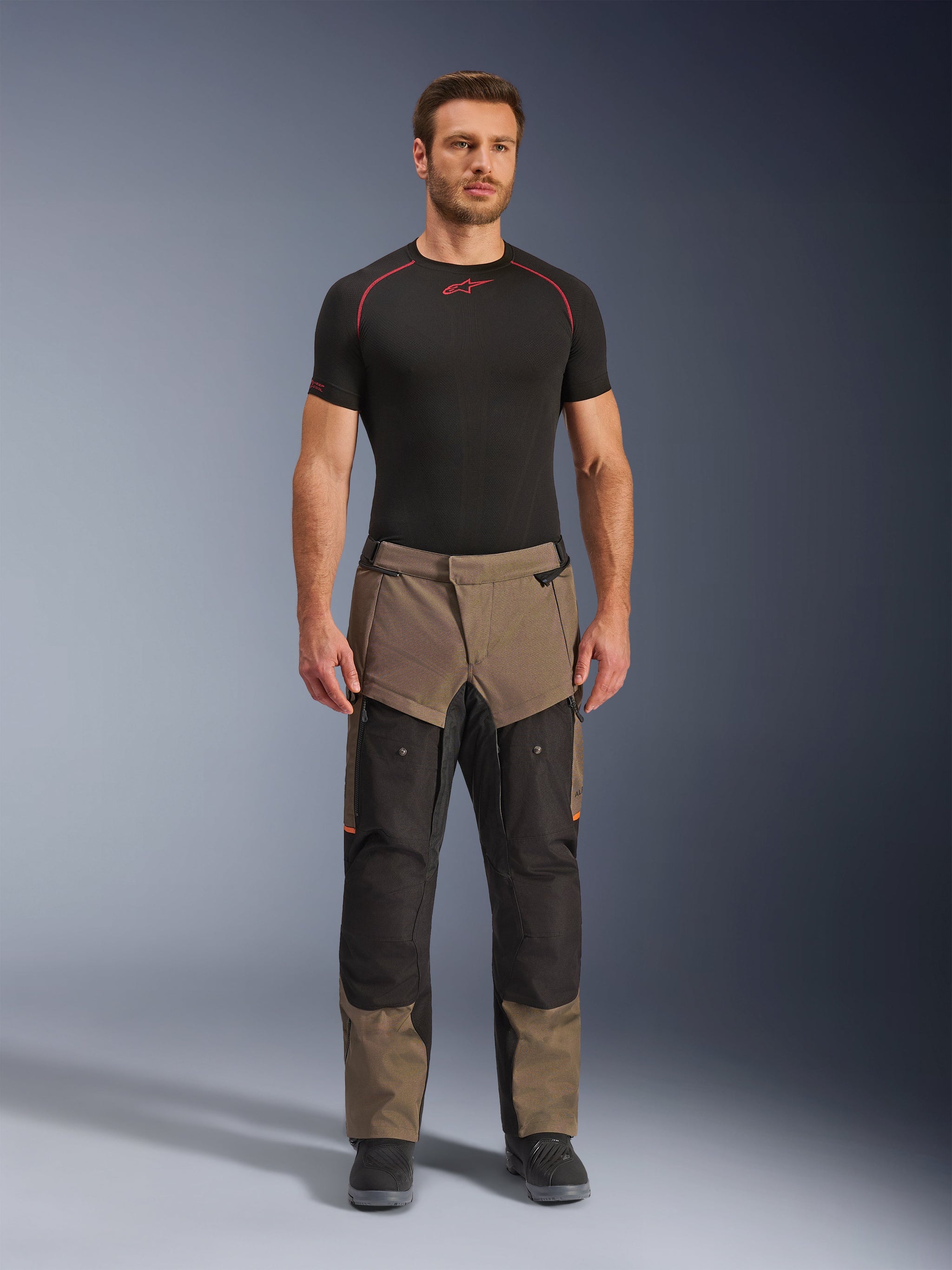 Alpinestars Cusco Drystar® broek, Walnut Black, adventure touring motorbroek, gedragen door een mannelijk model met een zwarte basislaag en zwarte laarzen, voorzien van walnootkleurige en zwarte panelen, oranje accenten en directe ventilatieritsen op de bovenbenen.