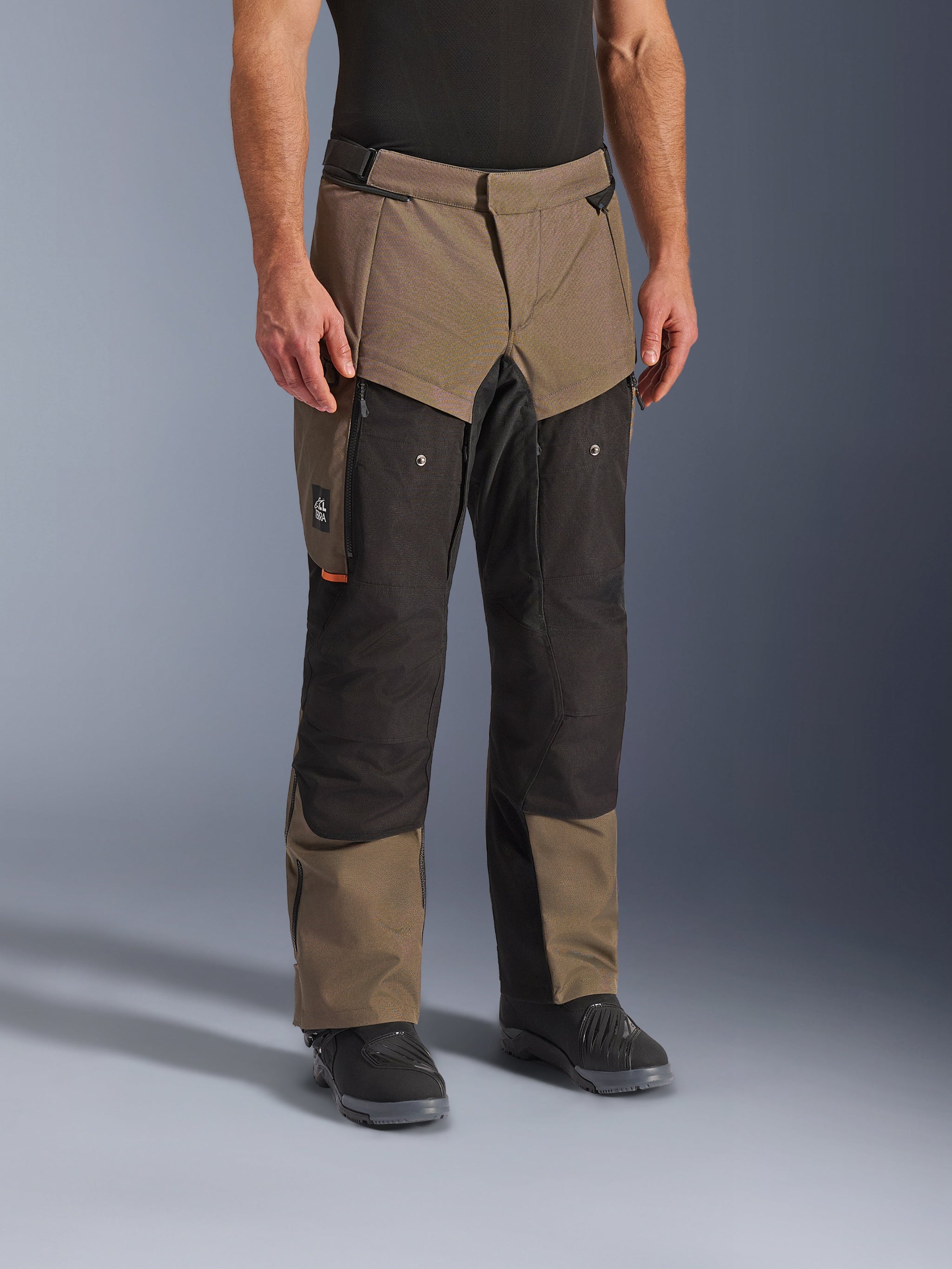 Alpinestars Cusco Drystar® broek, adventure motorbroek, Walnut Black, getoond op een model met zwarte laarzen, voorzien van een bruin en zwart multi-paneel ontwerp met ritszakken en versterkte knieën voor touring