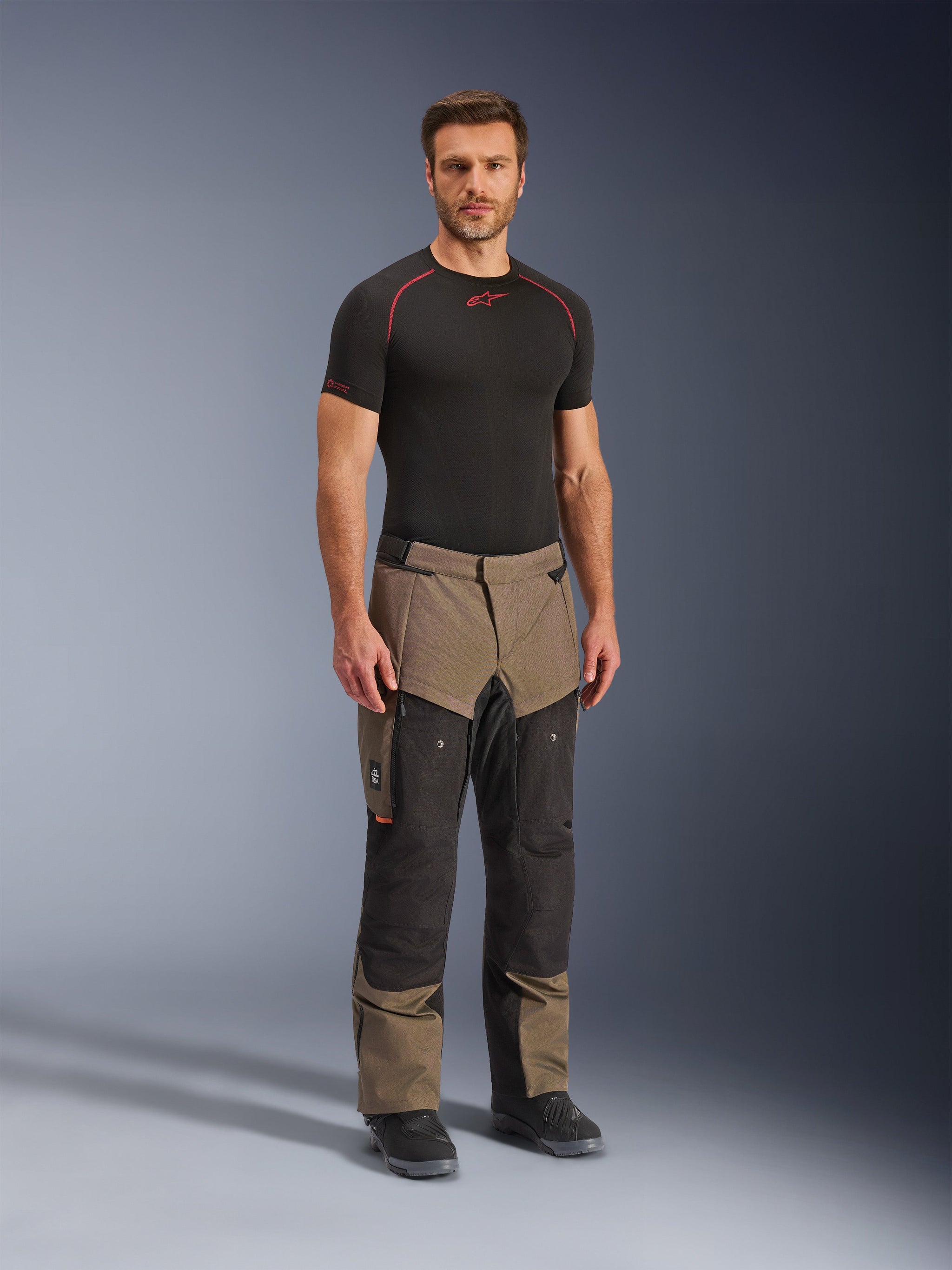Alpinestars Cusco Drystar® broek, adventure motorbroek, Walnut en zwart, gedragen door een model met een zwarte basislaag en laarzen, met grote cargozakken op de bovenbenen, ventilatieritsen en een robuust tweekleurig ontwerp.
