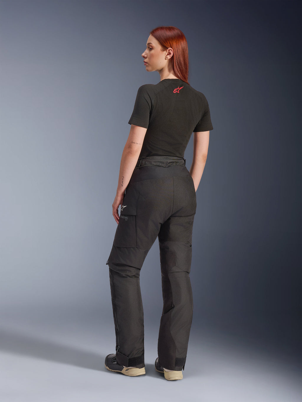 Alpinestars Stella Andes V4 Drystar® broek, zwarte adventure motorbroek, afgebeeld vanuit een zij-achteraanzicht op een model, gecombineerd met een zwart T-shirt en laarzen, voorzien van cargozakken met witte logo's en een duurzame textielconstructie.