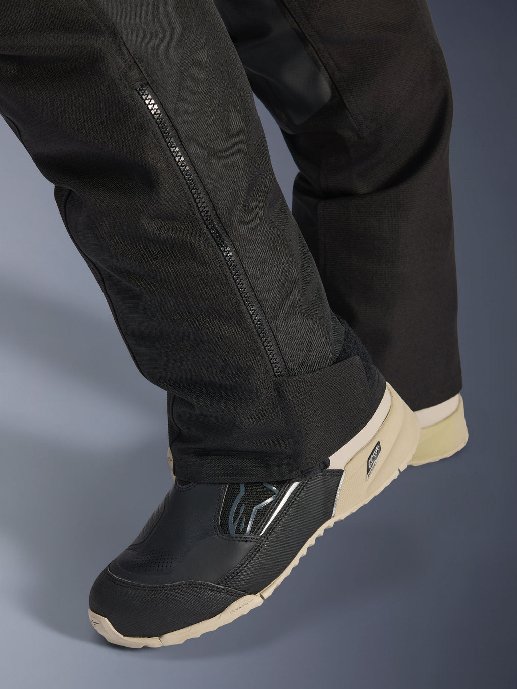 Alpinestars Stella Andes V4 Drystar® broek, zwart, close-up van het onderbeen met zijrits voor aanpassing van de laars, duurzaam textielmateriaal, gedragen met zwart en beige motorlaarzen, functioneel ontwerp klaar voor avontuur
