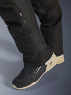 Alpinestars Stella Andes V4 Drystar® broek, zwart, close-up van het onderbeen met zijrits voor aanpassing van de laars, duurzaam textielmateriaal, gedragen met zwart en beige motorlaarzen, functioneel ontwerp klaar voor avontuur