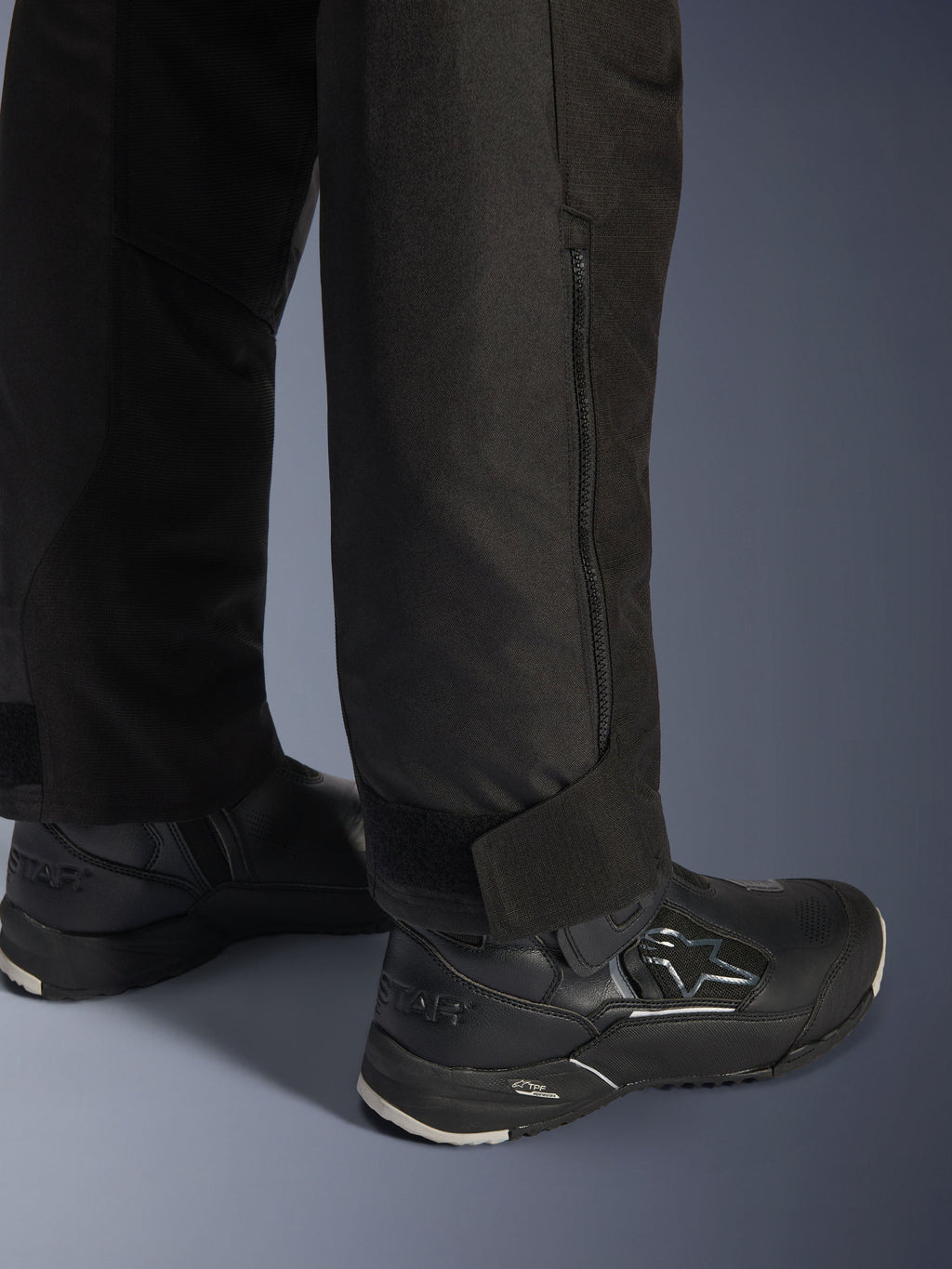Alpinestars Stella Andes V4 Drystar® broek, zwart, close-up van de onderkant van de broekspijp en de boord, voorzien van een ritssluiting aan de zijkant en een verstelbare klittenbandsluiting bij de enkel, gedragen met zwarte Alpinestars motorlaarzen.