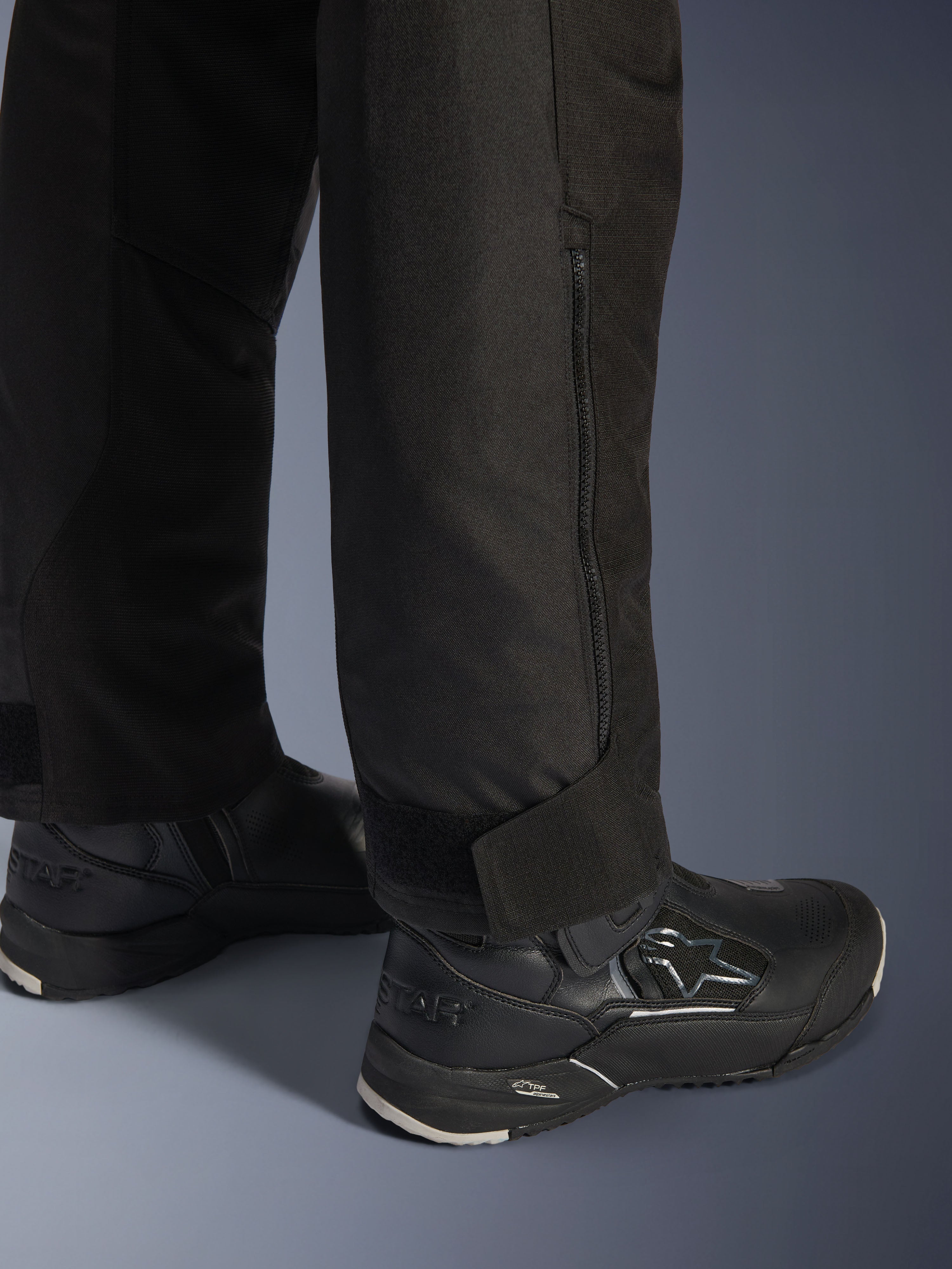 Alpinestars Stella Andes V4 Drystar® broek, zwart, close-up van de onderkant van de broekspijp en de boord, voorzien van een ritssluiting aan de zijkant en een verstelbare klittenbandsluiting bij de enkel, gedragen met zwarte Alpinestars motorlaarzen.